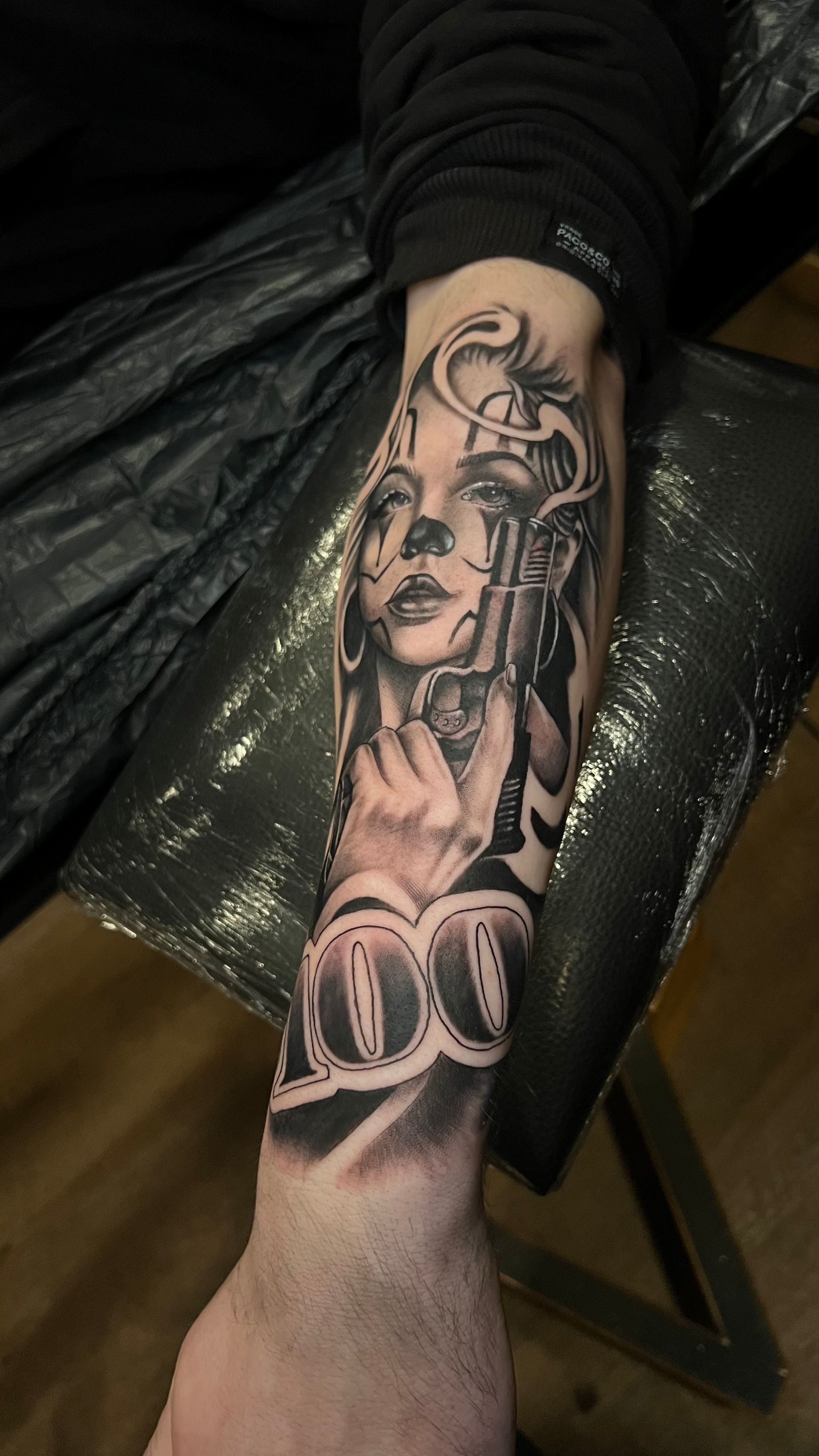 A black-and-grey tattoo on a forearm featuring a figure with clown makeup holding a handgun, with 
