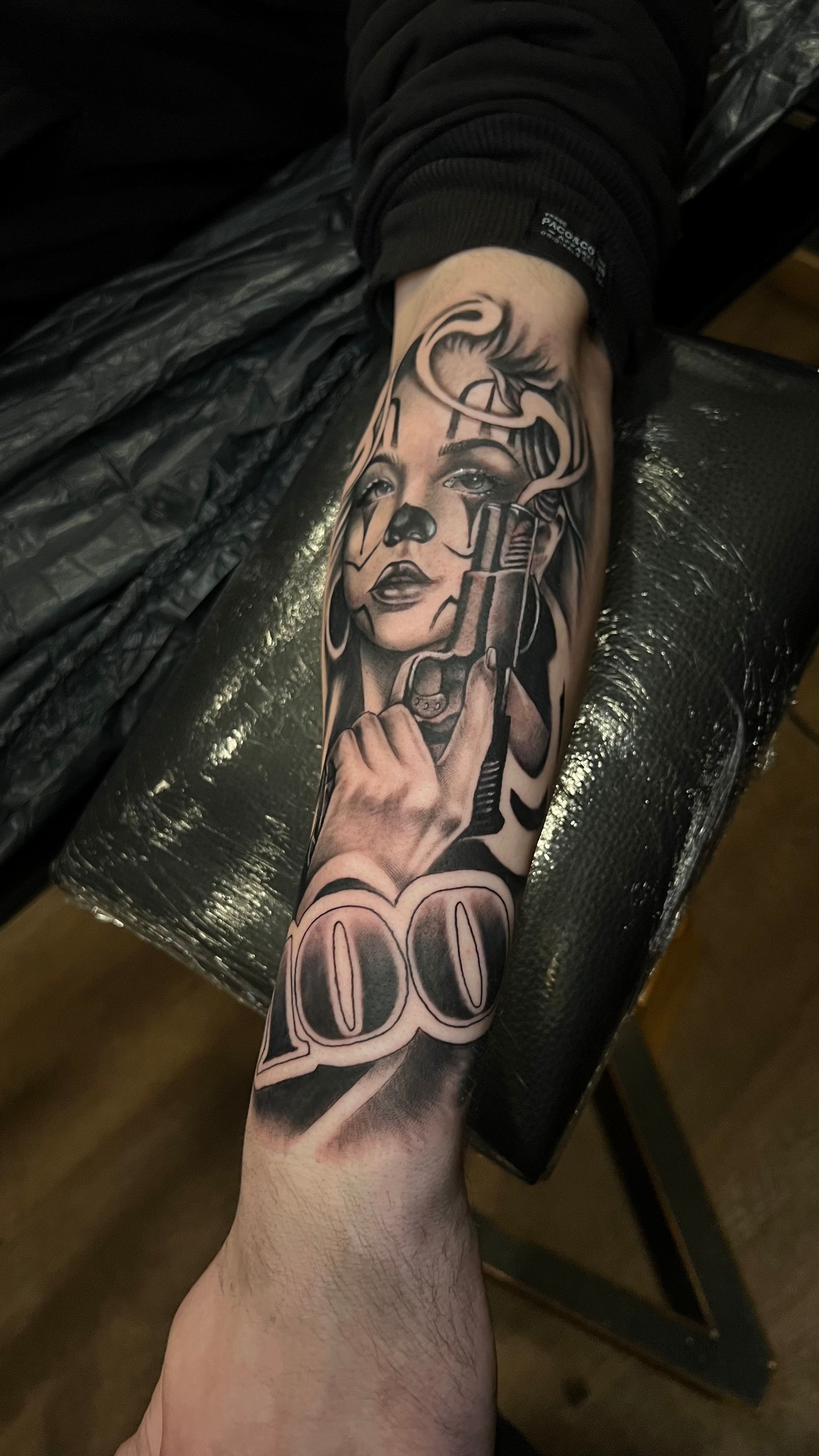 A forearm tattoo of a person with clown-style face paint holding a handgun, featuring the number 100 below it.