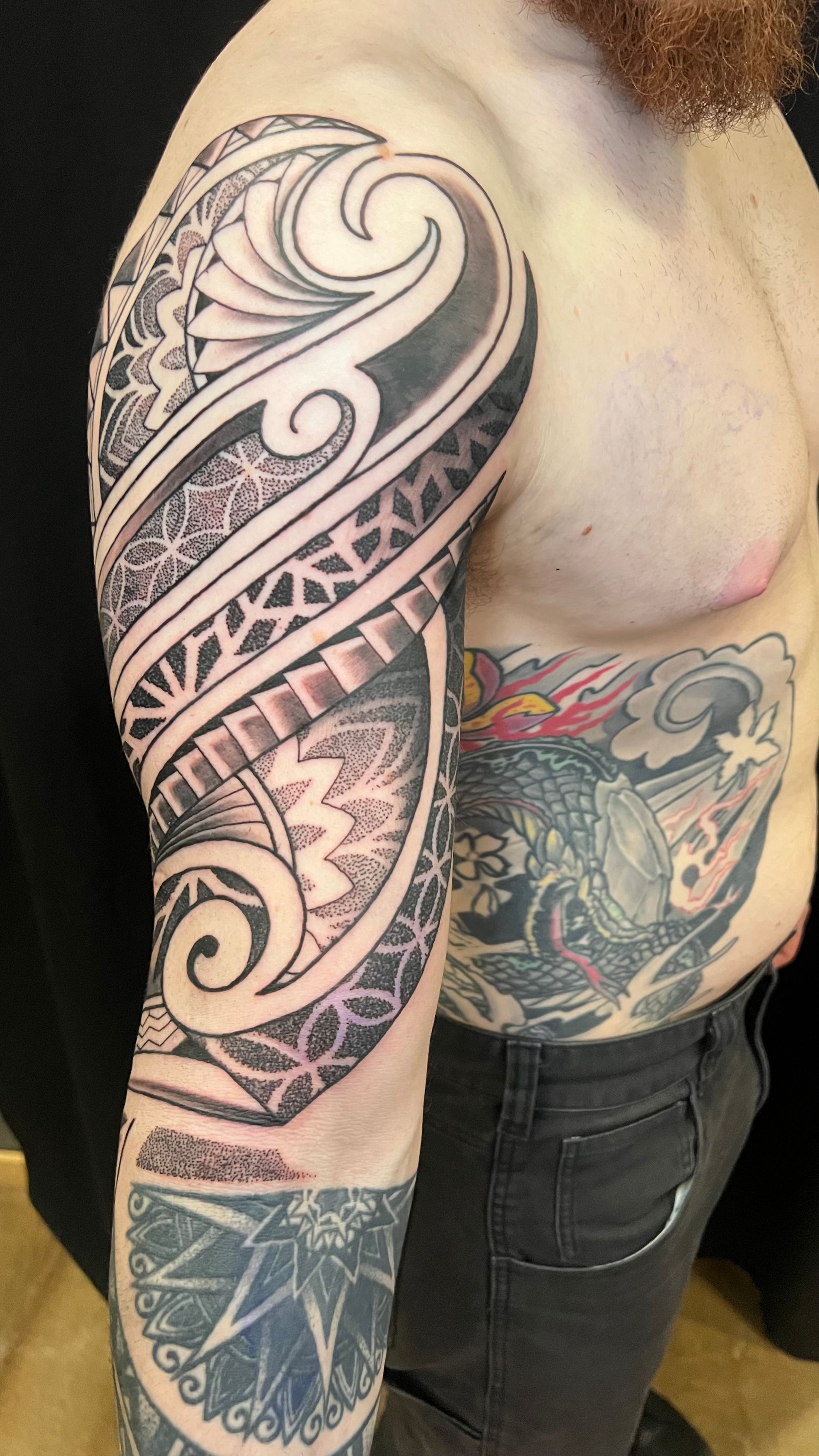 A person with elaborate tribal-style blackwork tattoos on their upper arm and additional tattoos on their torso and forearm.