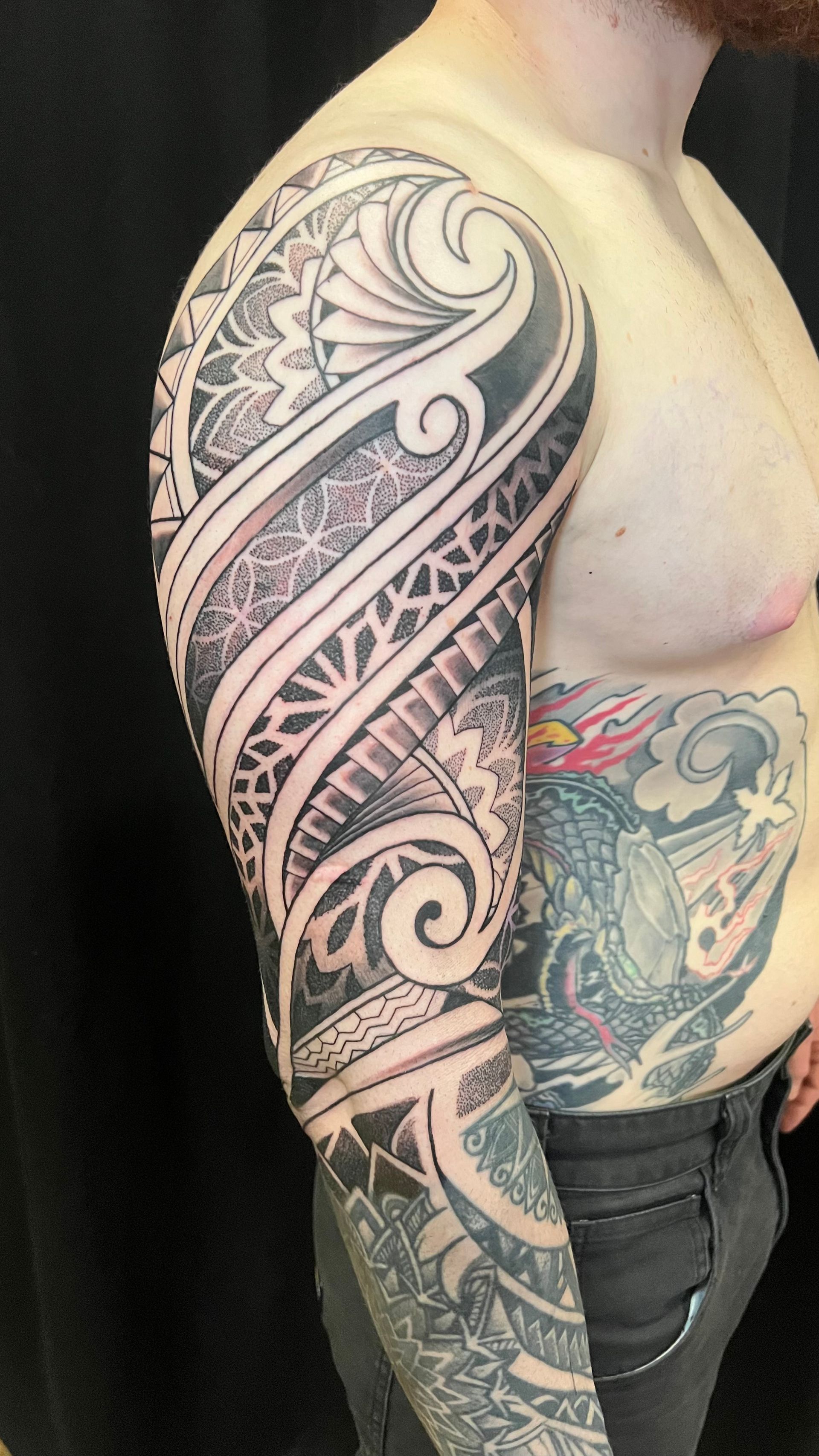 A person's arm displaying a detailed black and grey Polynesian-style tribal tattoo covering their shoulder and sleeve.