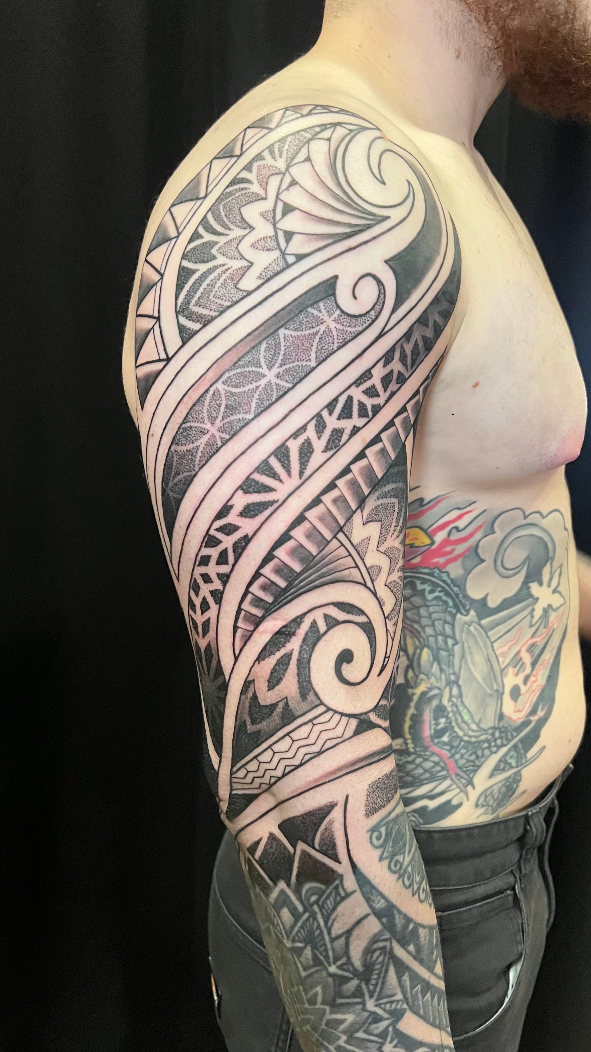 A side view of a person’s arm featuring an intricate black-ink sleeve tattoo with bold geometric and swirling patterns.