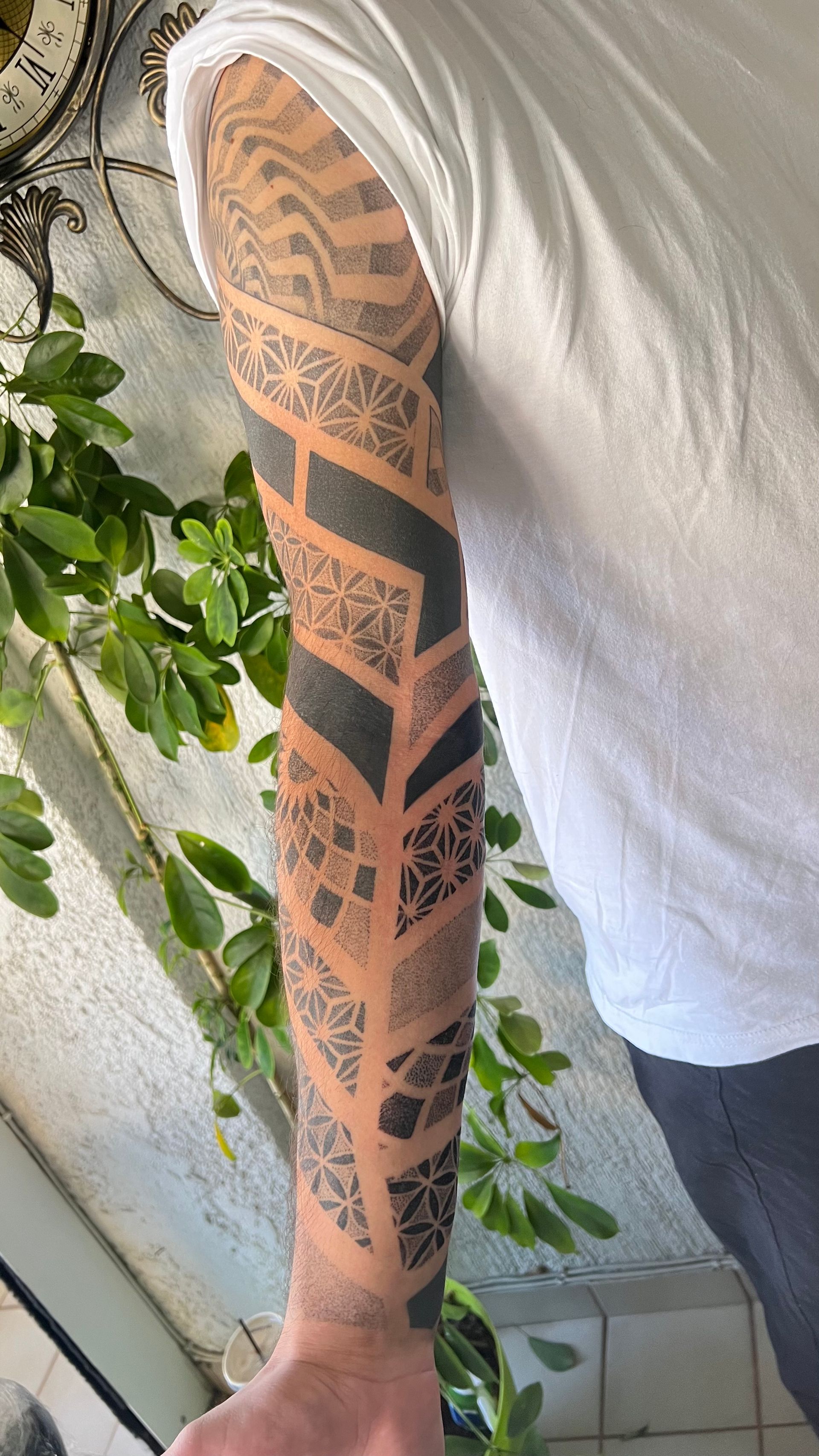 A person wearing a white t-shirt displays an arm covered in a dark, geometric Polynesian-style tattoo sleeve.