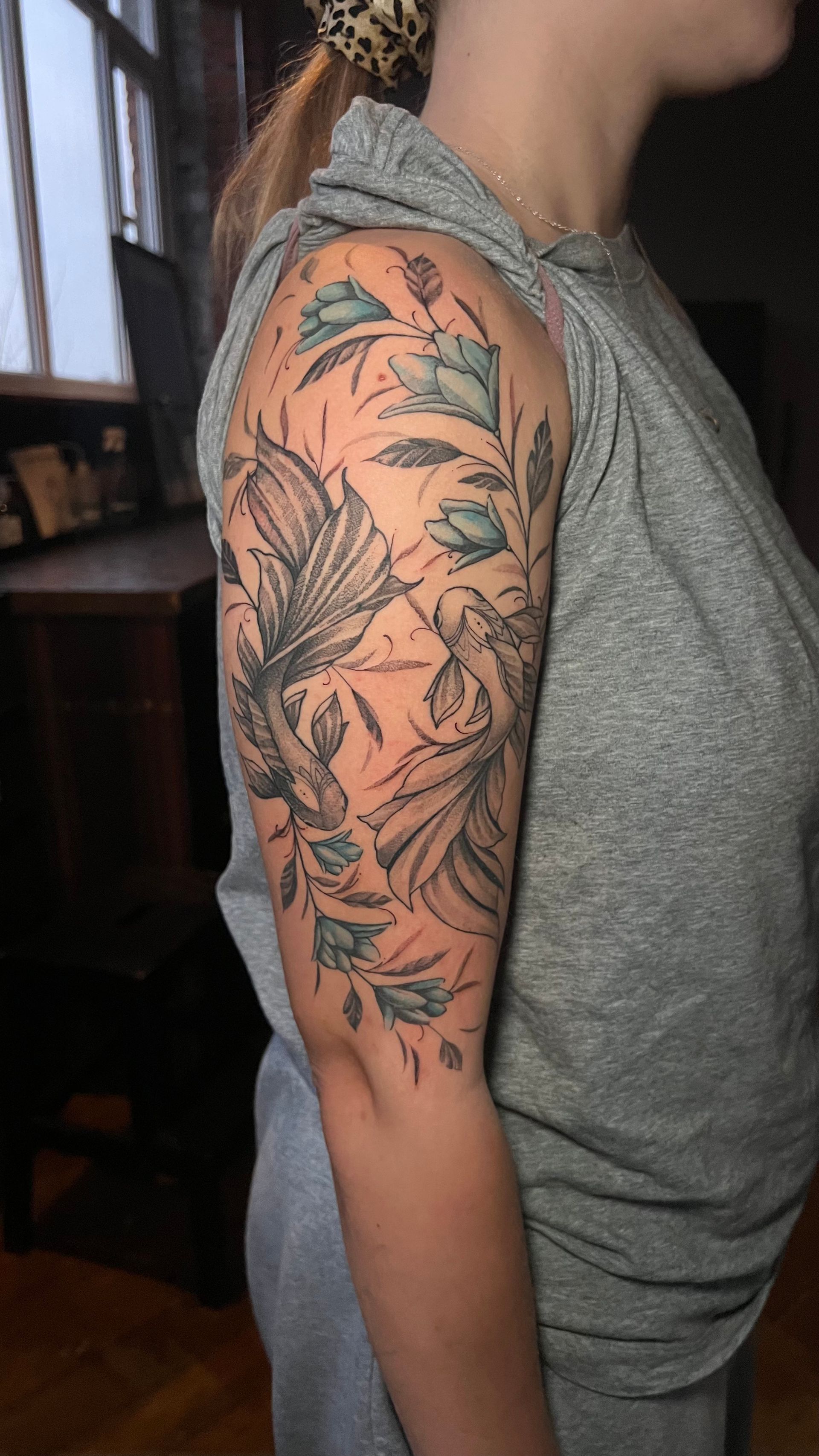 Side profile of an upper arm displaying a tattoo featuring two fish surrounded by flowing blue-tinted botanical elements.