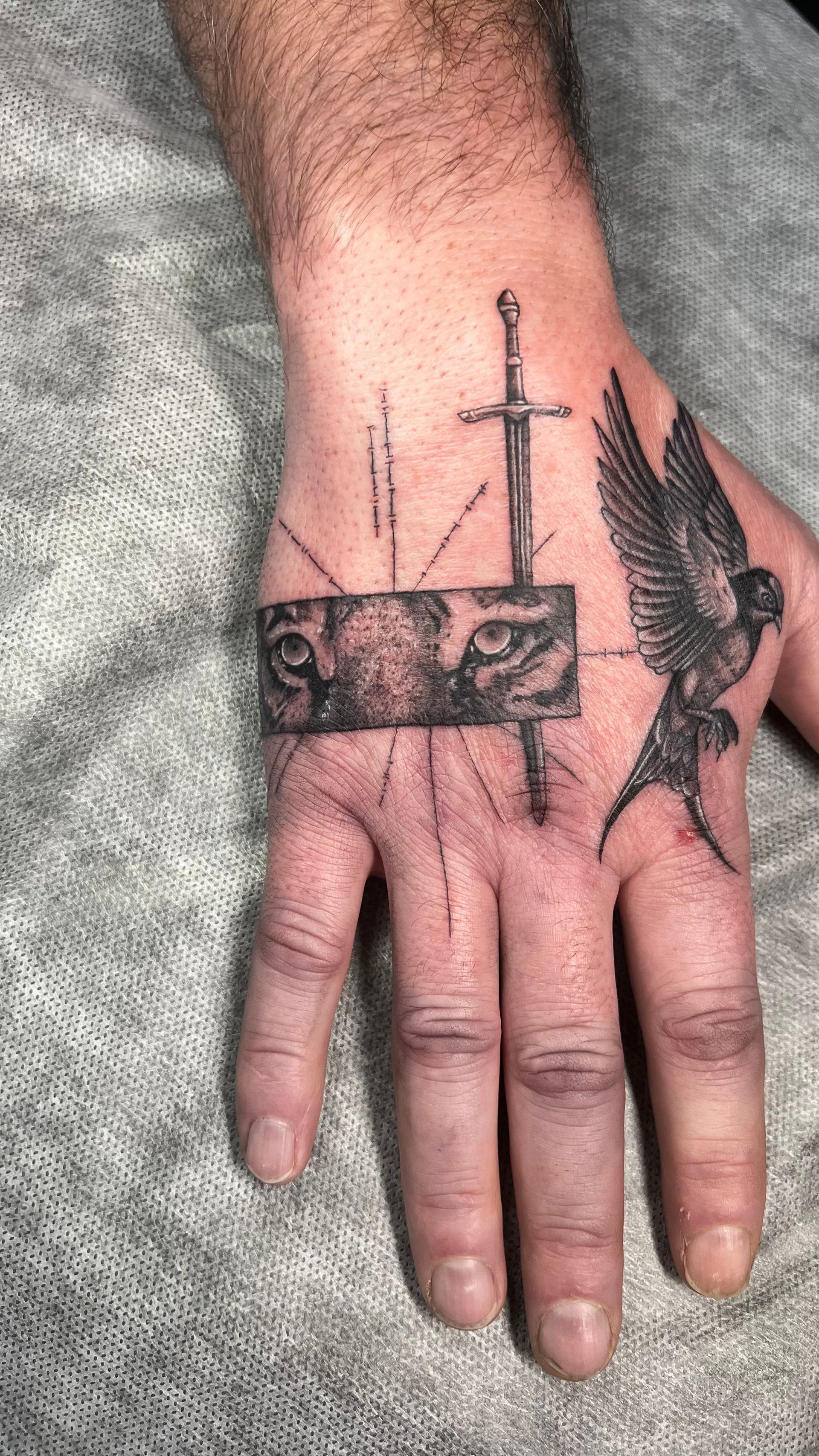 Black ink tattoo on a hand featuring a rectangular tiger eye design, a vertical sword, and a bird in flight.