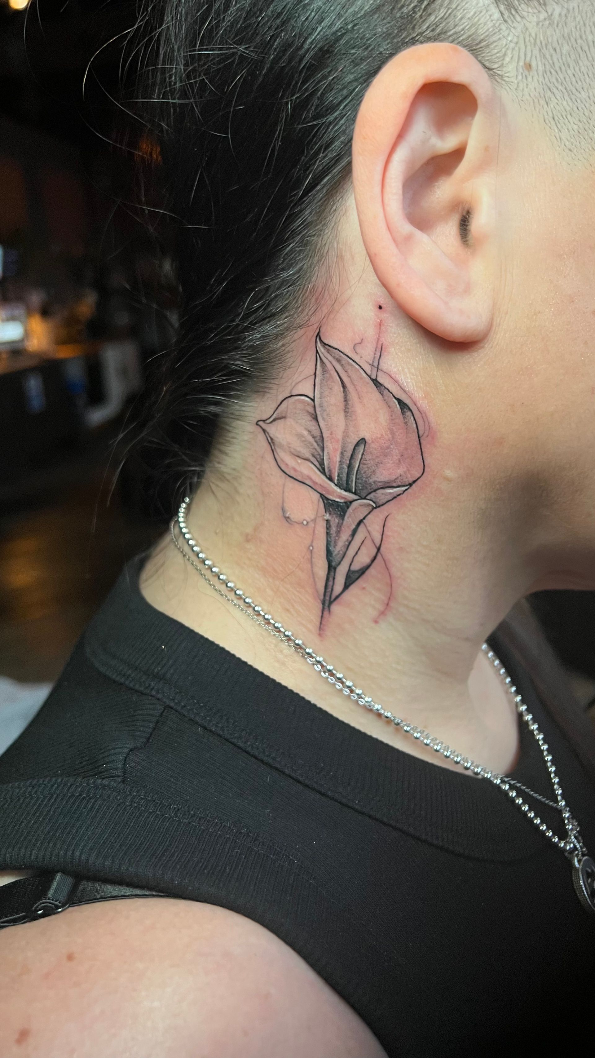 A delicate, fine-line black ink tattoo of a calla lily flower on the side of a person’s neck, just below the ear.