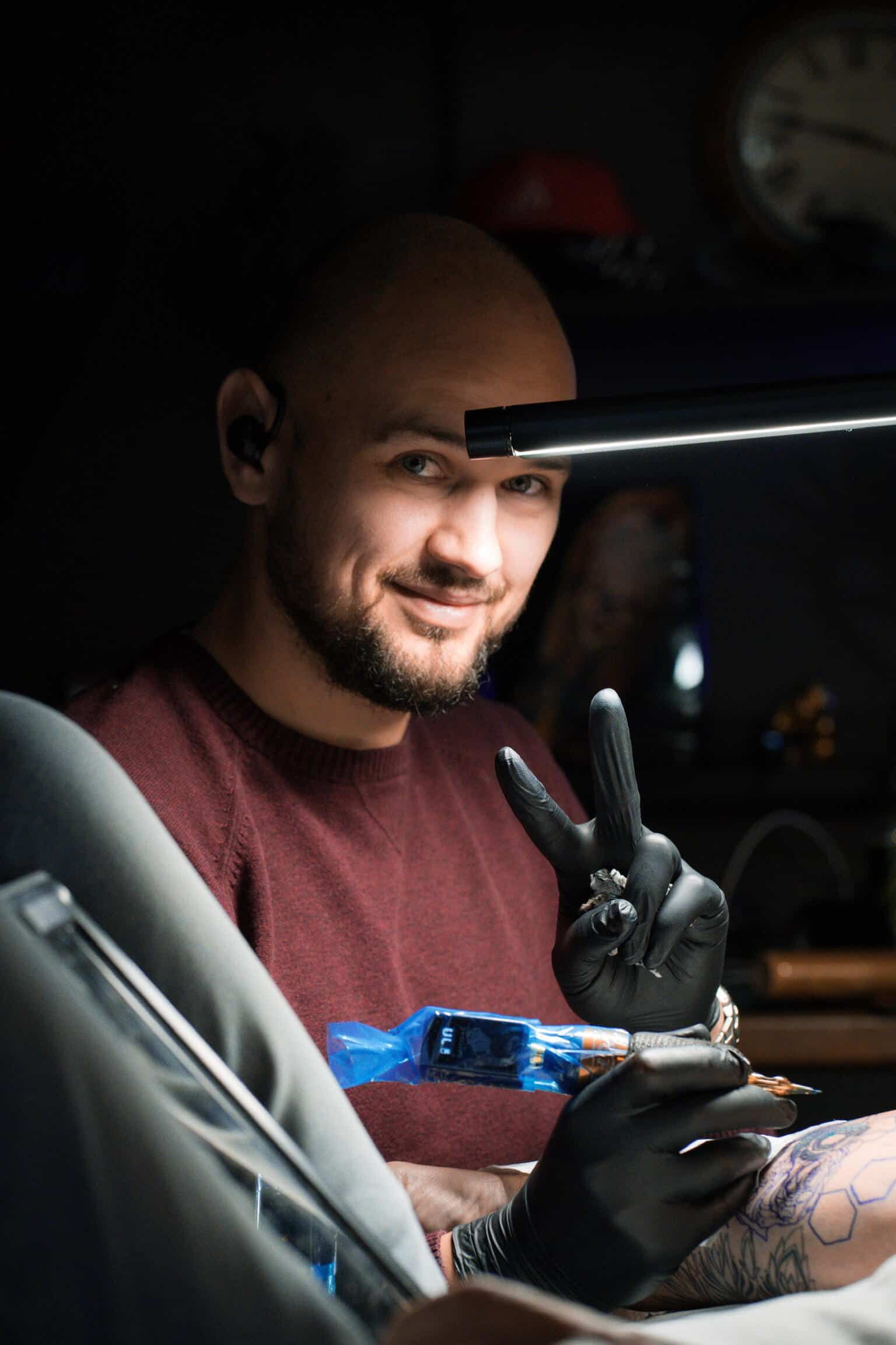 A smiling tattoo artist in a maroon sweater and black gloves makes a peace sign while working on a tattoo.