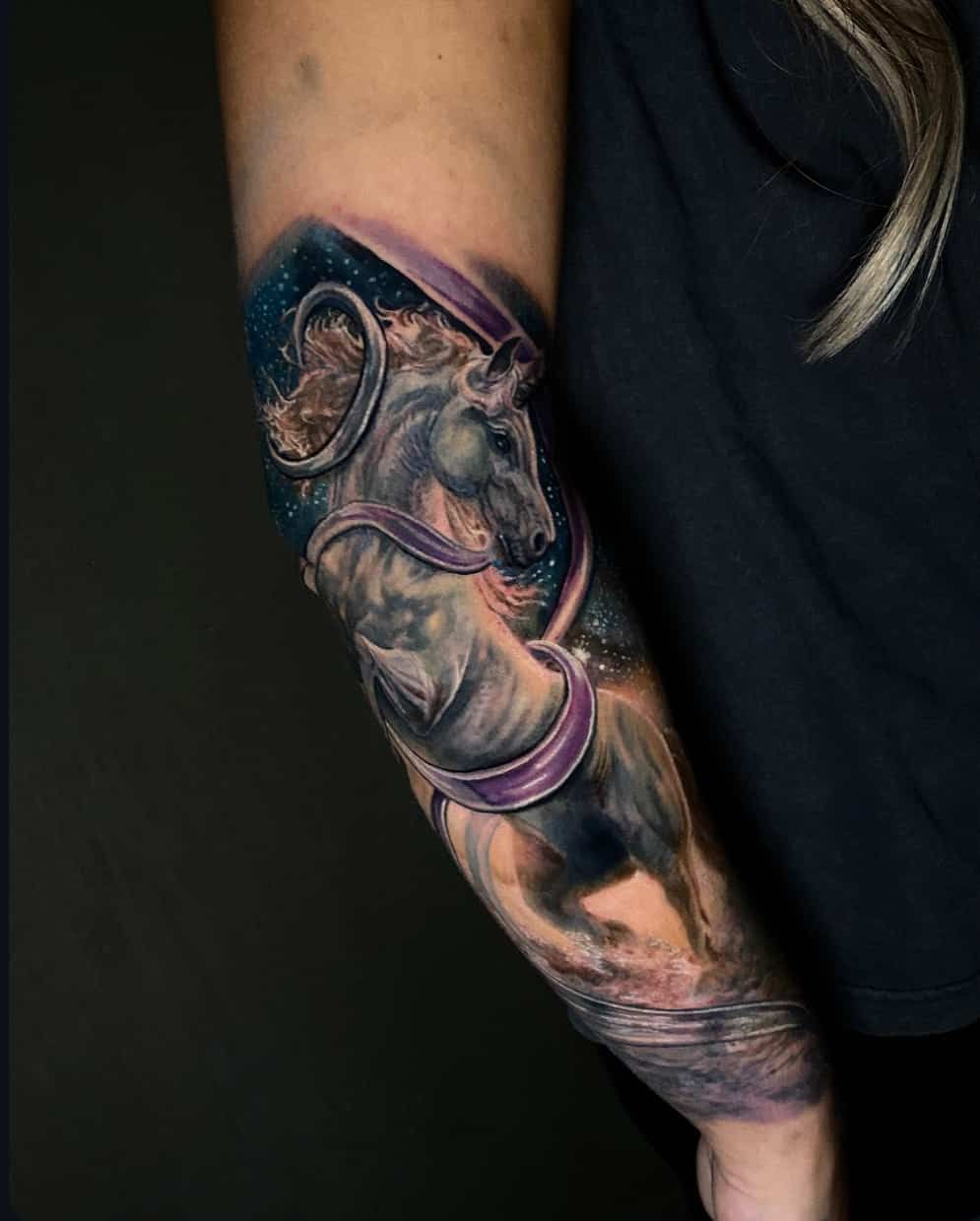 A detailed forearm tattoo of a unicorn entwined in purple ribbons against a dark, starry night sky background.