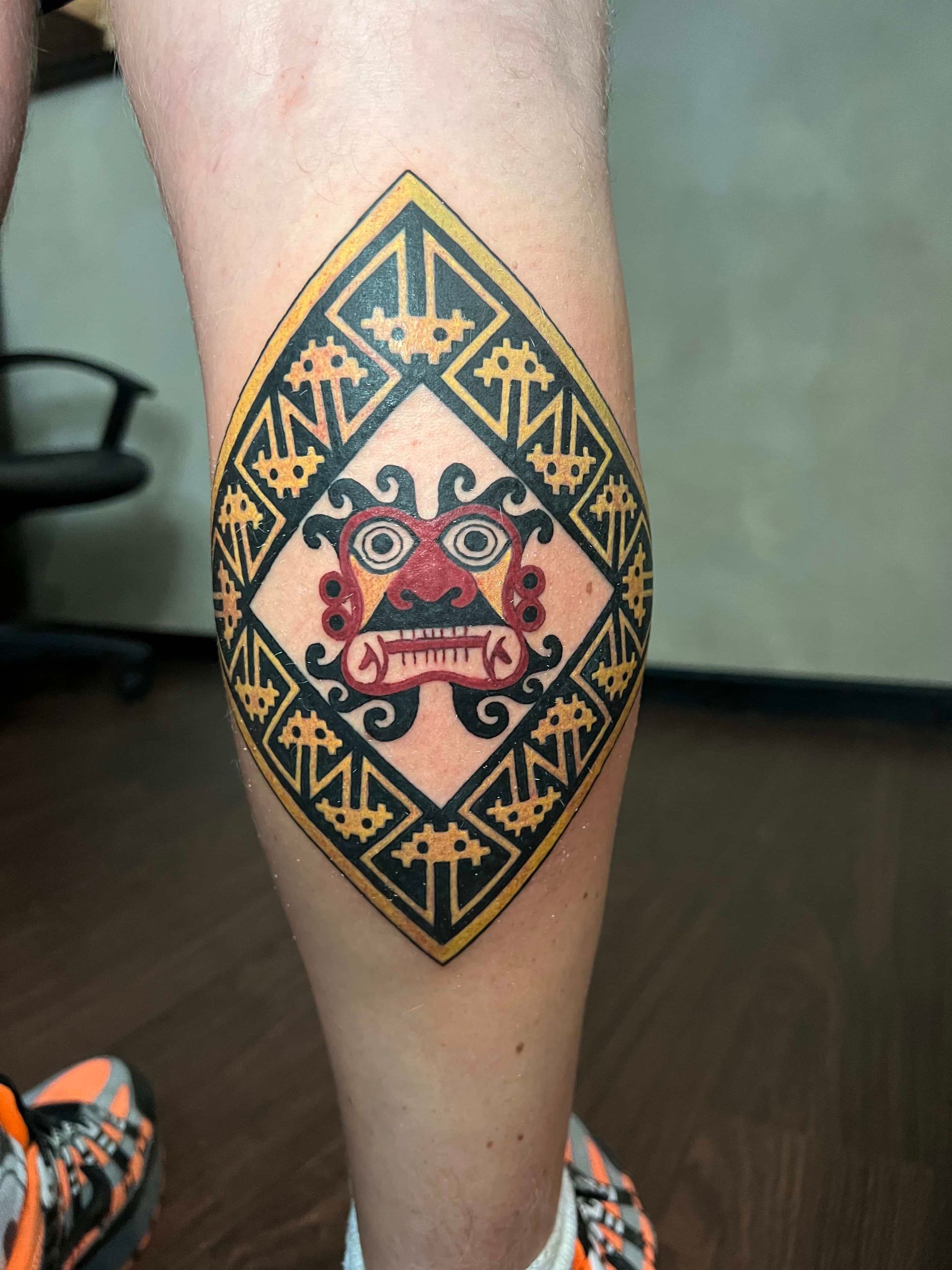 A diamond-shaped tattoo on a person's calf featuring a stylized, colorful deity mask surrounded by intricate black patterns.