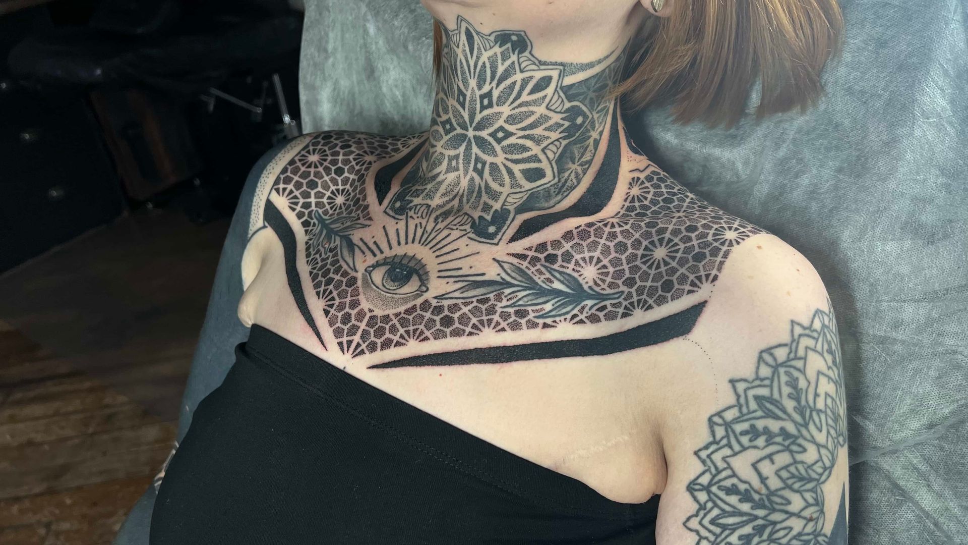 A person wearing an intricate, black-ink chest and neck tattoo featuring a central eye, sun rays, and geometric patterns.