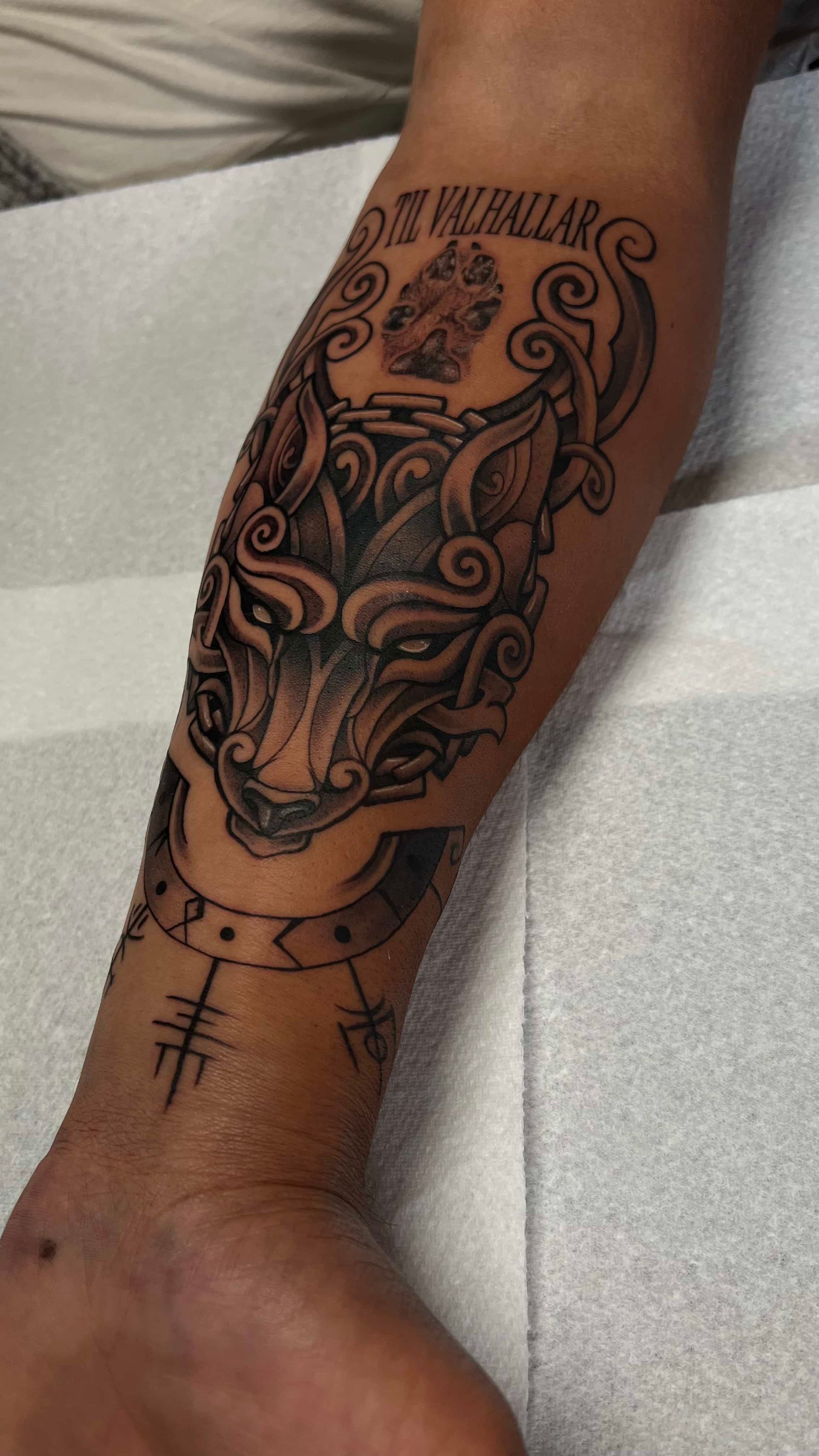 Black-ink tattoo on a forearm featuring a stylized wolf face, decorative knots, runic symbols, and text reading 