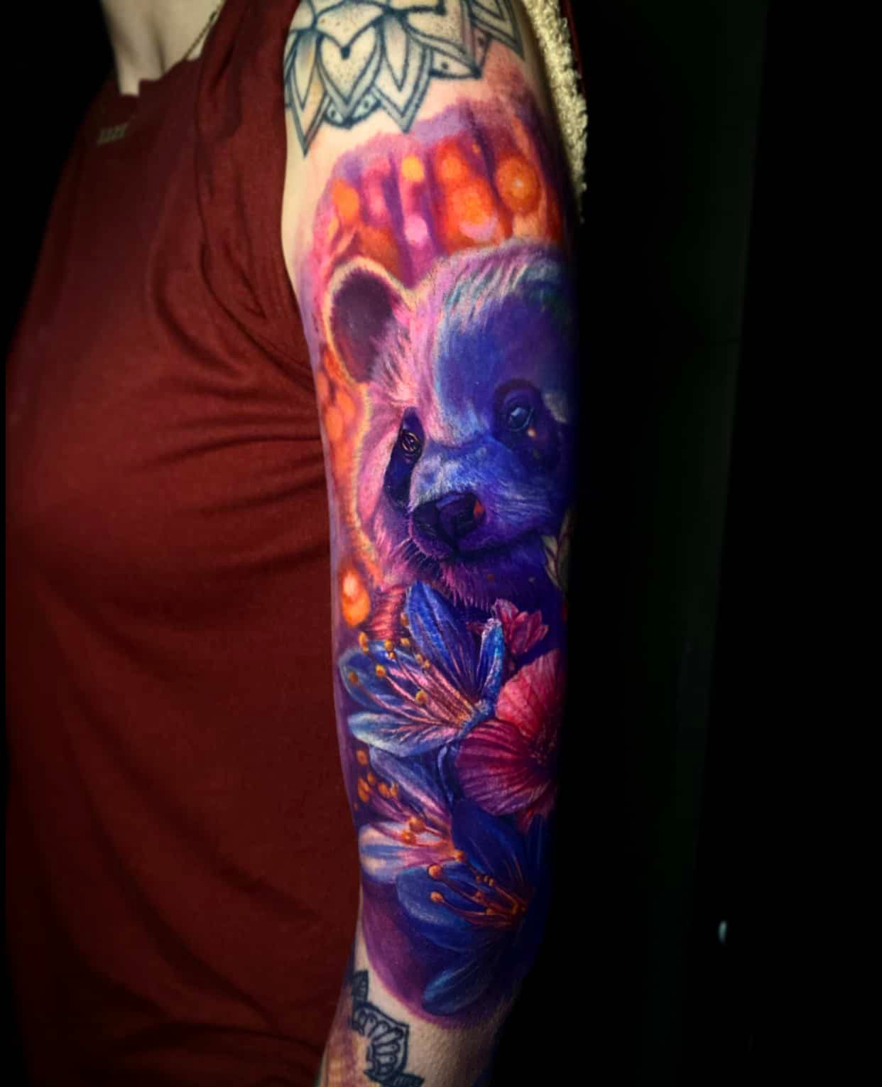 A vibrant tattoo on an arm featuring a panda face surrounded by blue and purple flowers against a glowing orange backdrop.