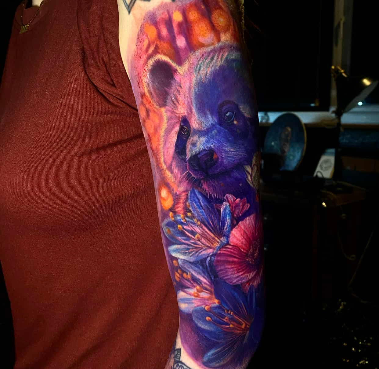 A colorful, vibrant tattoo on an arm featuring a panda surrounded by blue and magenta flowers with an orange background.