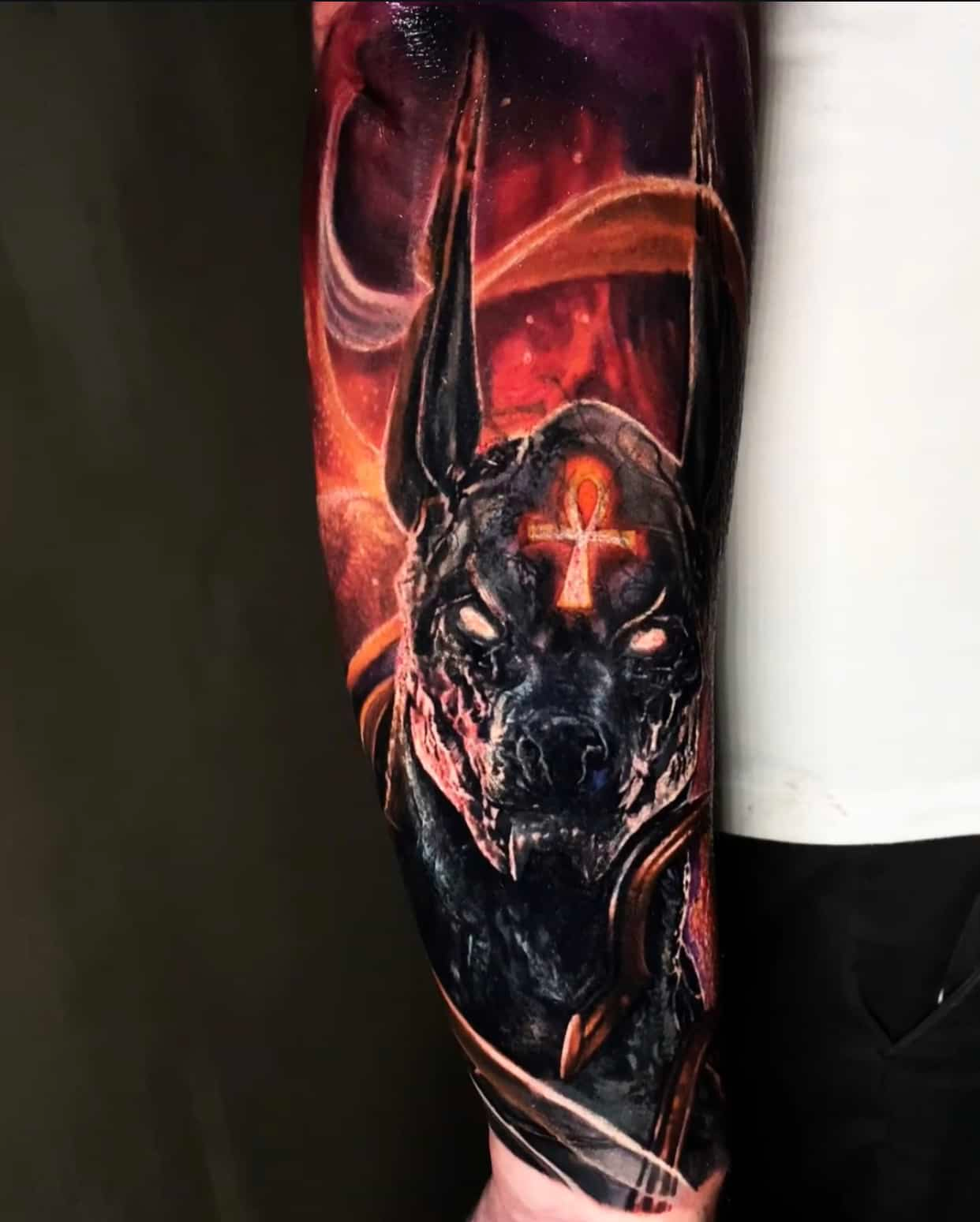 A tattoo on a forearm depicting the Egyptian god Anubis, rendered in dark tones with an glowing orange ankh and backdrop.
