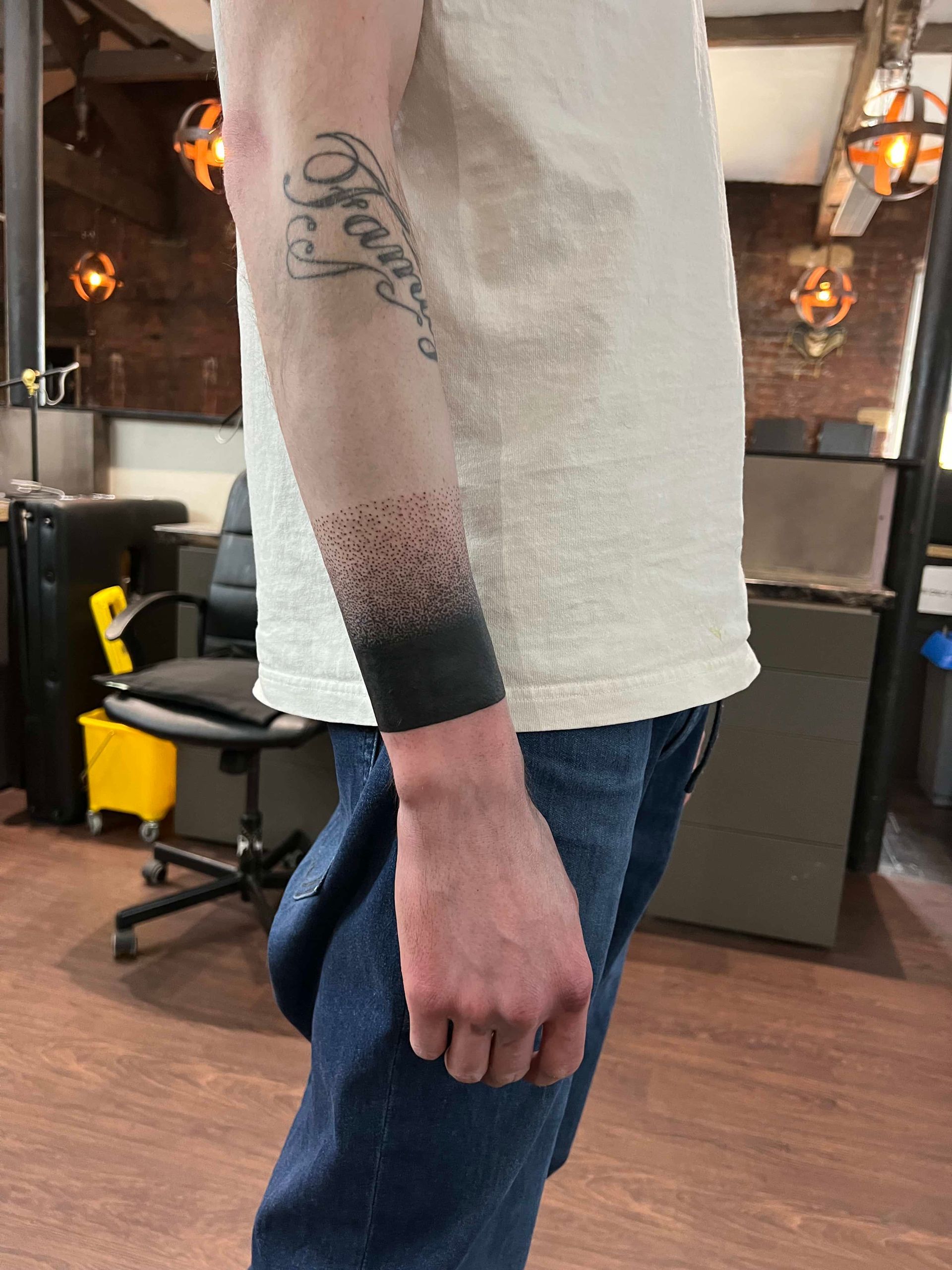 A person’s forearm with a dark, solid band tattoo that fades into a dotwork stippling pattern near the elbow.