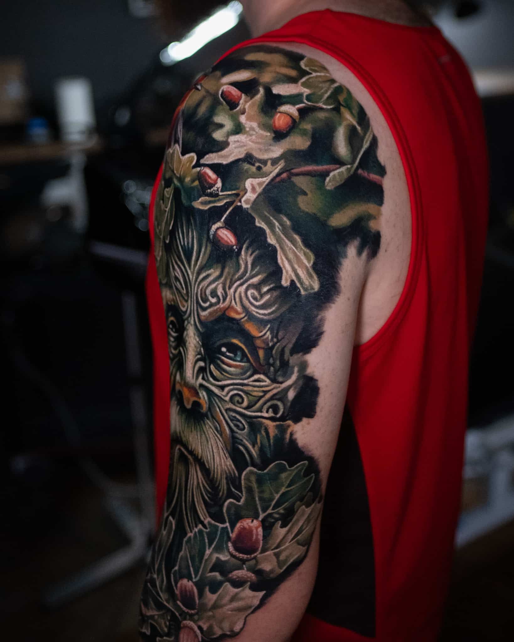 A shoulder tattoo featuring a detailed Green Man figure surrounded by oak leaves and acorns in shades of green and brown.