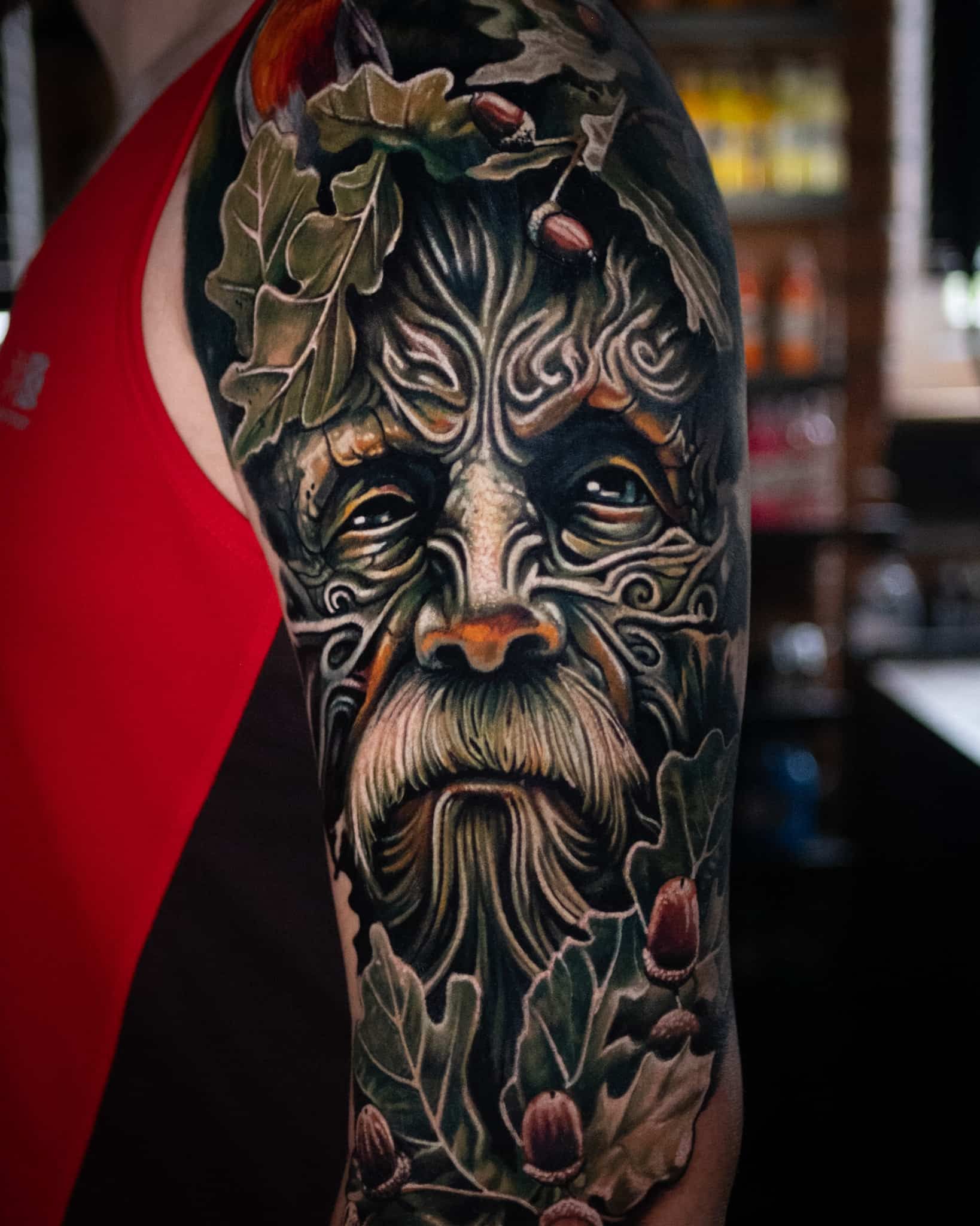Detailed upper arm tattoo of a Green Man surrounded by intricate oak leaves and acorns in shades of green and brown.