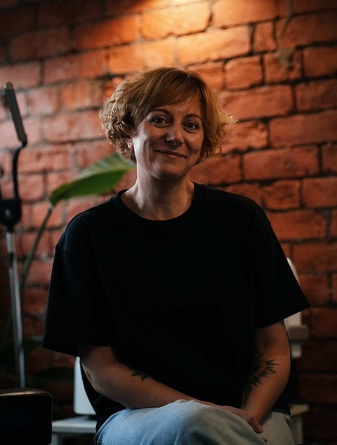A person with short, light hair smiling at the camera, wearing a black shirt against a textured red brick wall.