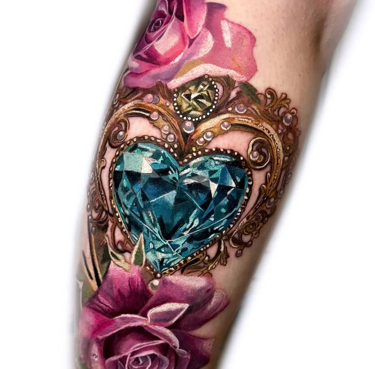 A realistic tattoo featuring a large, heart-shaped blue diamond set in ornate gold filigree, surrounded by pink roses.
