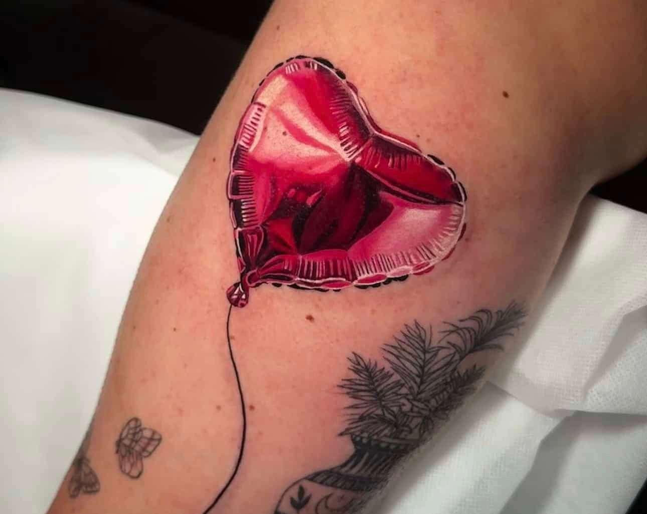 A realistic tattoo of a shiny, metallic red heart-shaped balloon with a thin string on skin, near a botanical tattoo.