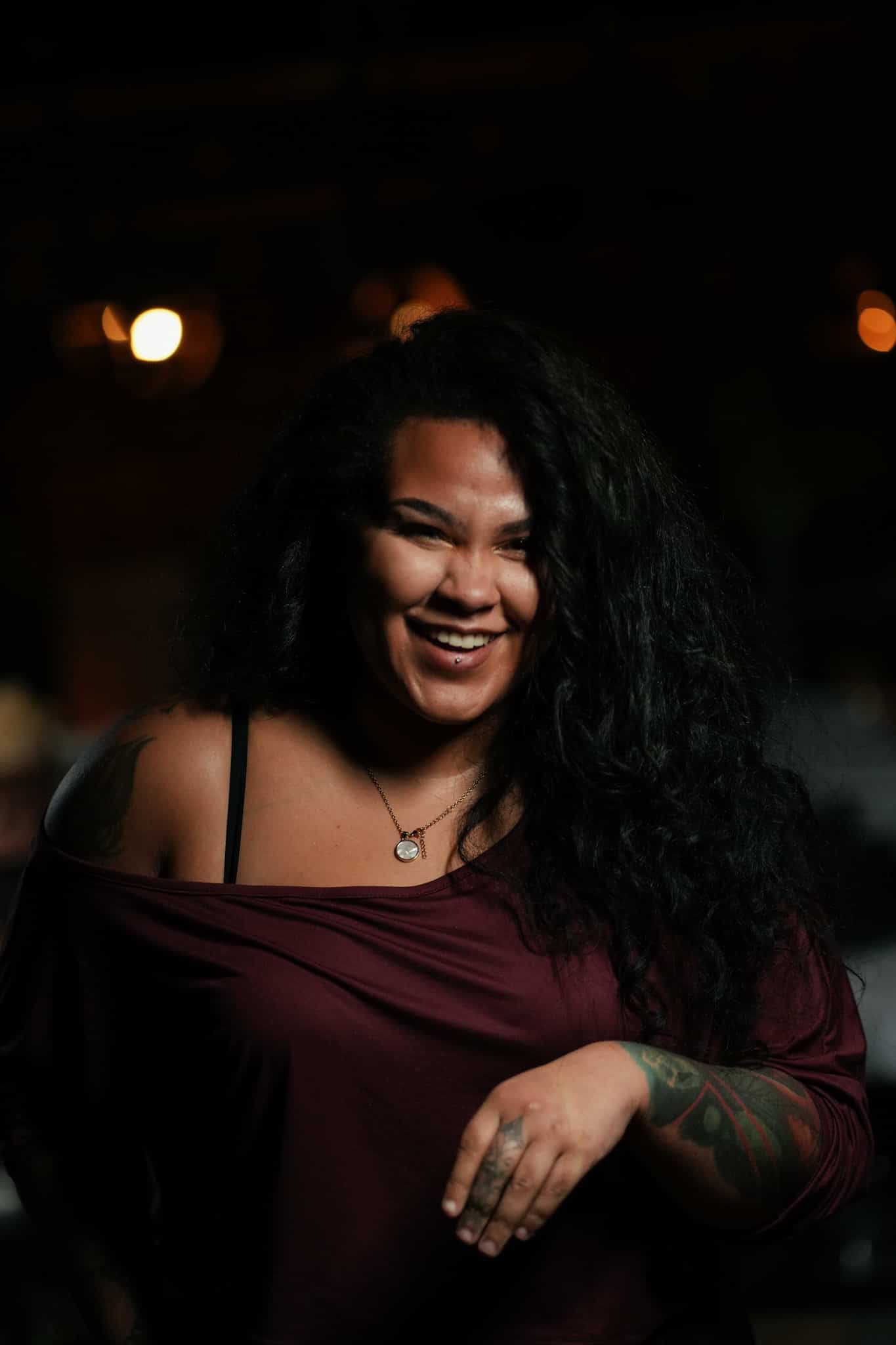 A smiling person with long dark wavy hair, wearing a dark red off-the-shoulder top and a necklace, posed in a dim setting.