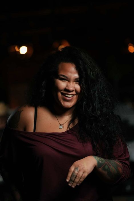 A smiling person with long dark wavy hair, wearing a dark red off-the-shoulder top and a necklace in a dimly lit setting.