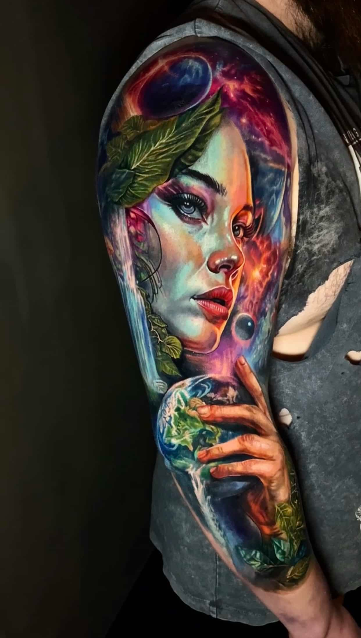 A vibrant, cosmic-themed tattoo on an upper arm featuring a face framed by foliage and a hand cradling a globe.