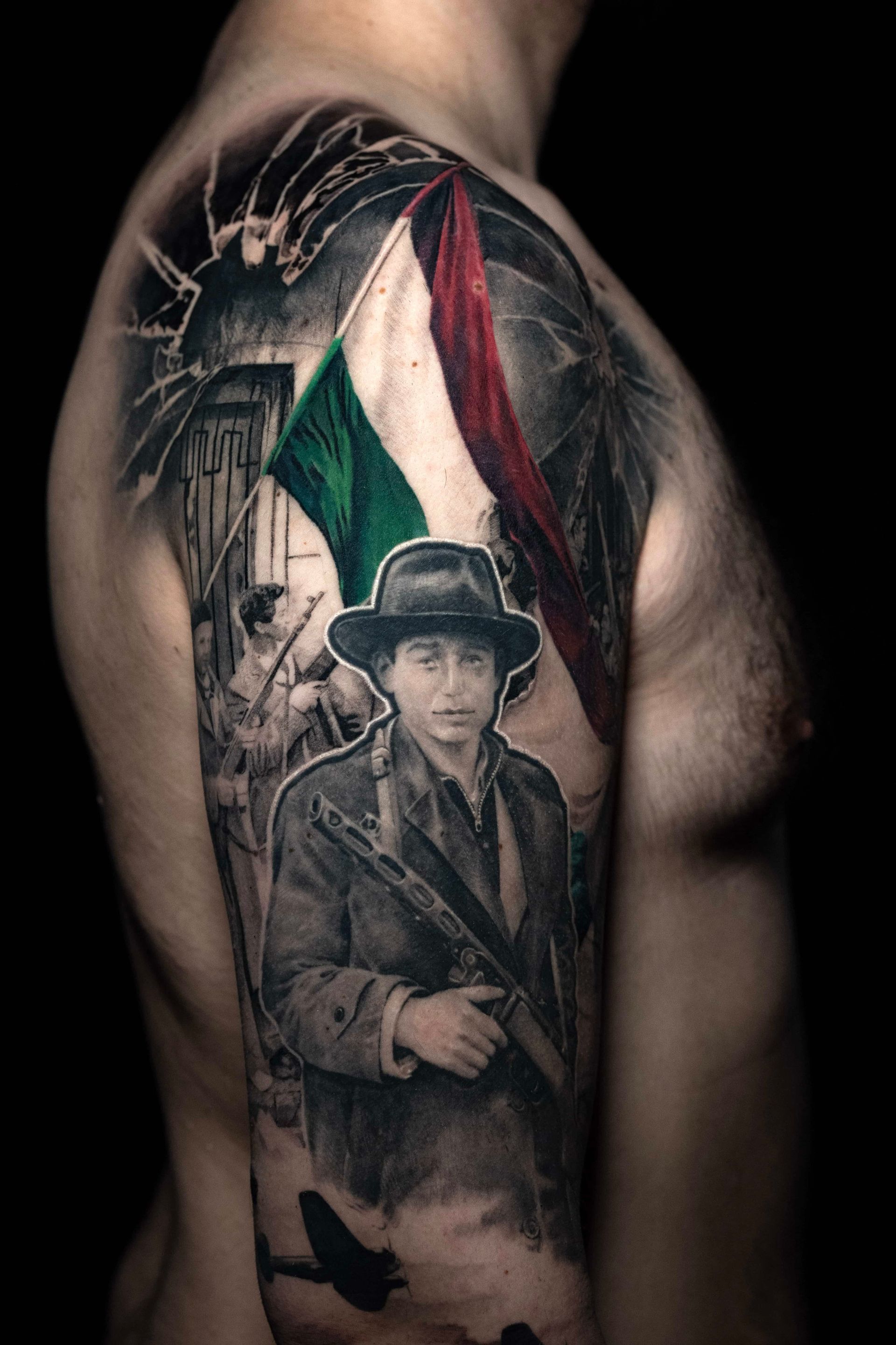 A detailed shoulder tattoo featuring a person in a hat holding a rifle beneath a colored Italian flag, with background art.