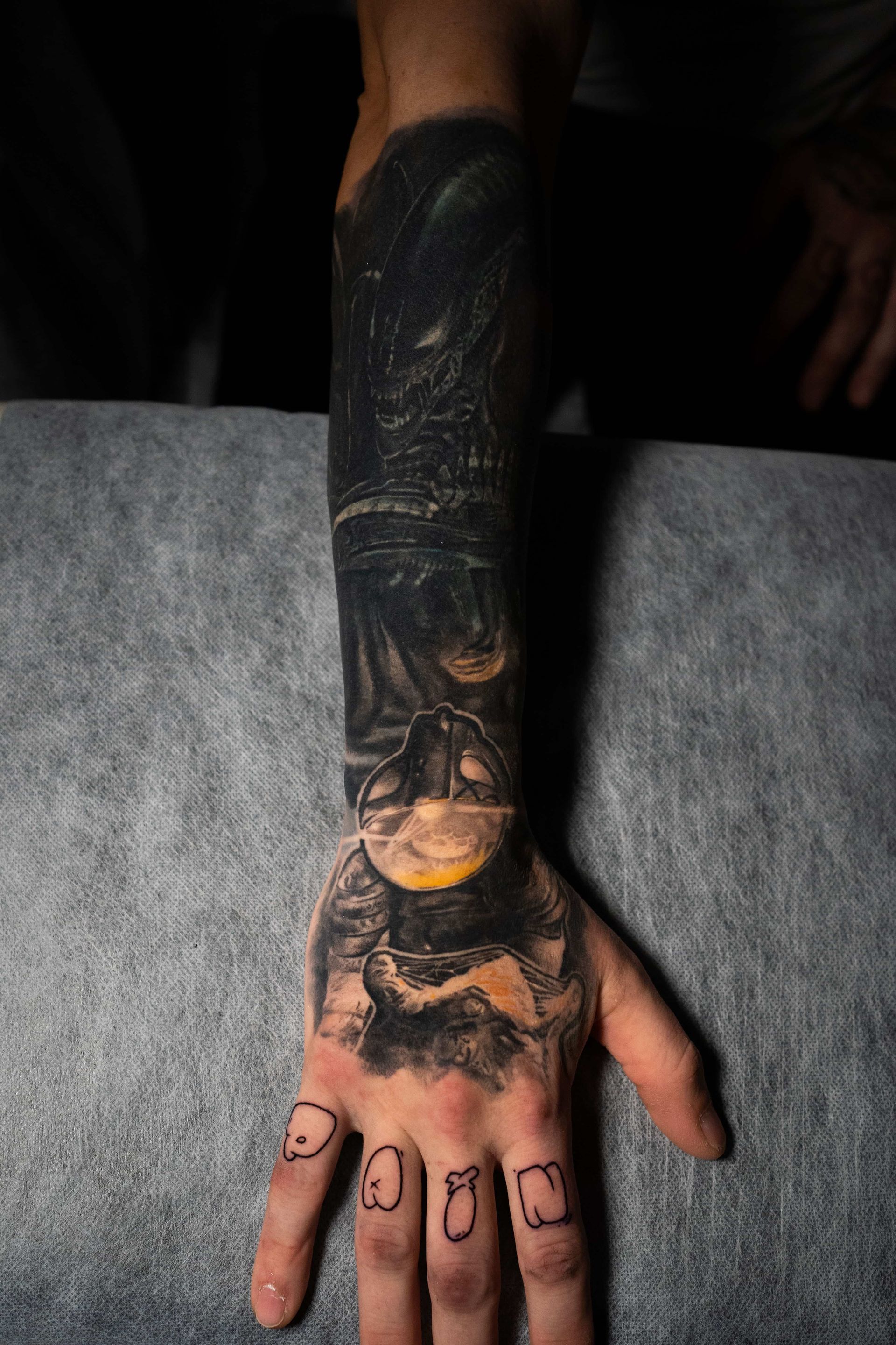 A forearm and hand featuring a detailed, dark-themed tattoo with a yellow accent, and simple line drawings on the fingers.