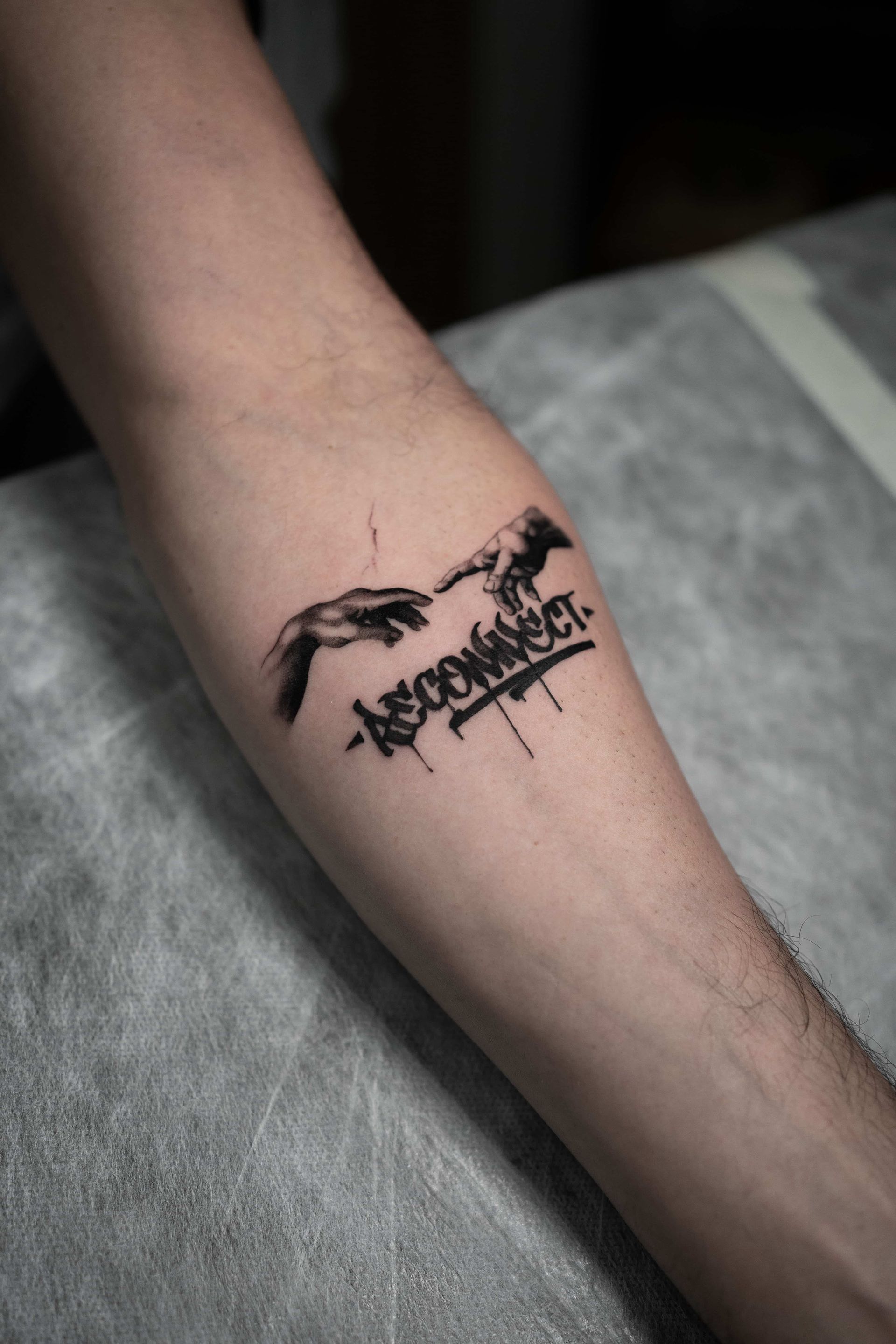 A tattoo on a forearm featuring the hands from The Creation of Adam with the word 