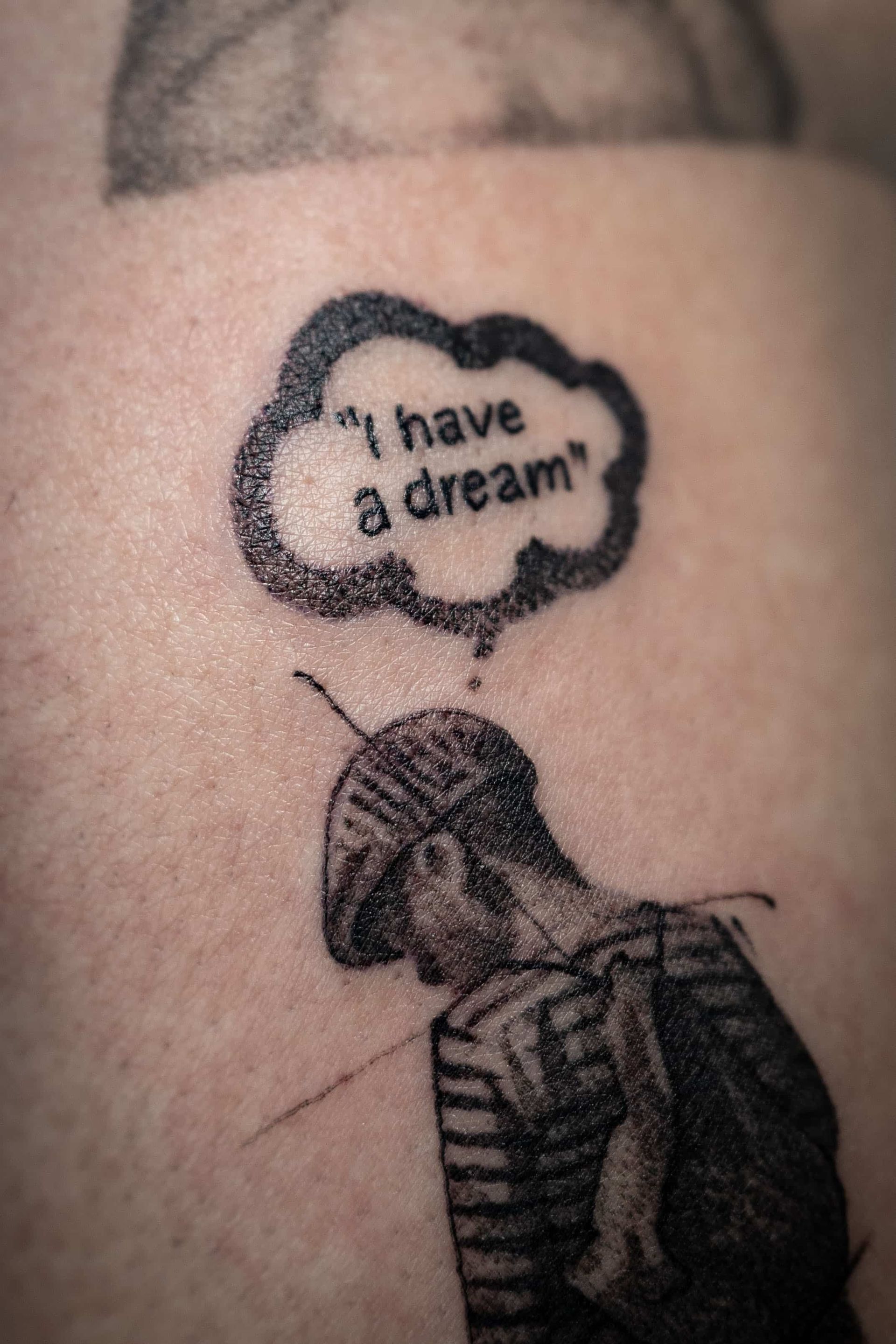 A black-and-white tattoo of a person in a striped shirt and beret looking up at a speech bubble reading 