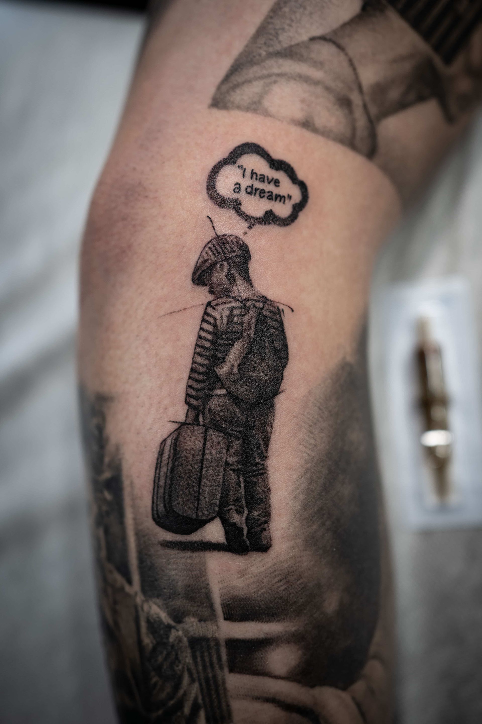 A small tattoo of a person wearing a cap and carrying a suitcase, with a thought bubble above reading 