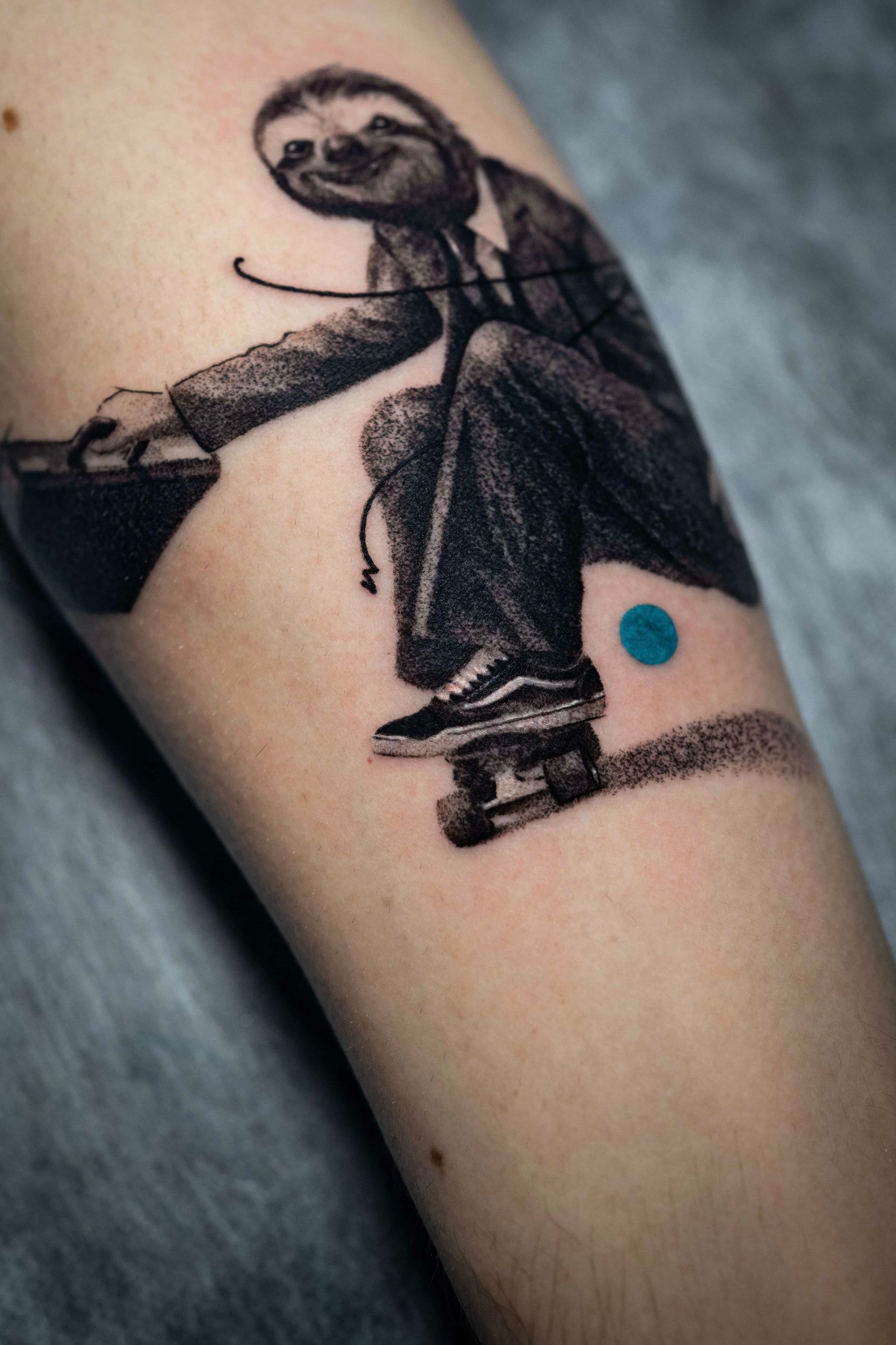 A detailed tattoo of a sloth wearing a suit and sneakers, skateboarding while carrying a briefcase, with a blue dot.