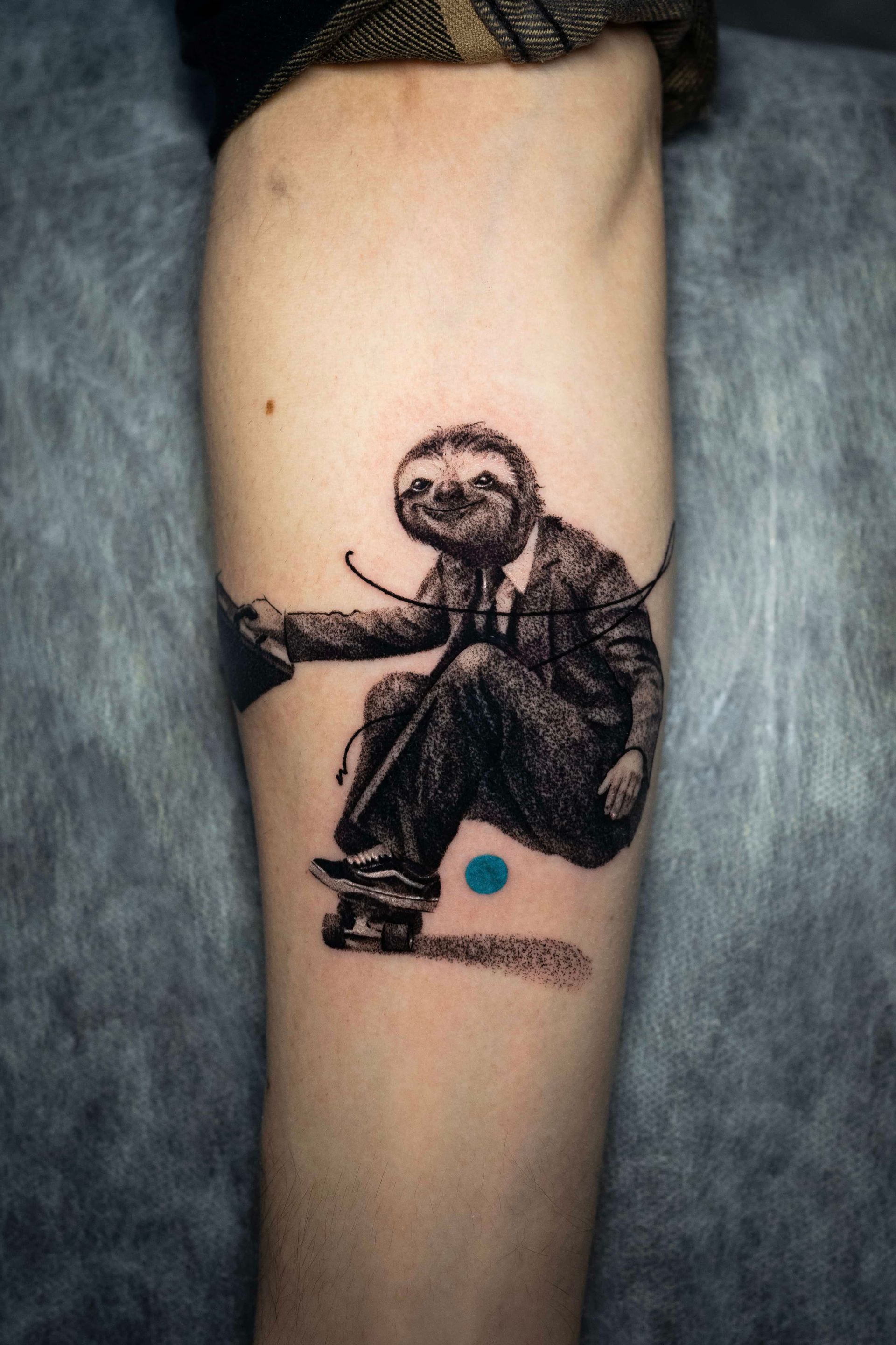 A black-ink tattoo of a sloth wearing a suit and hat, skateboarding, featuring a single, small blue dot below the board.