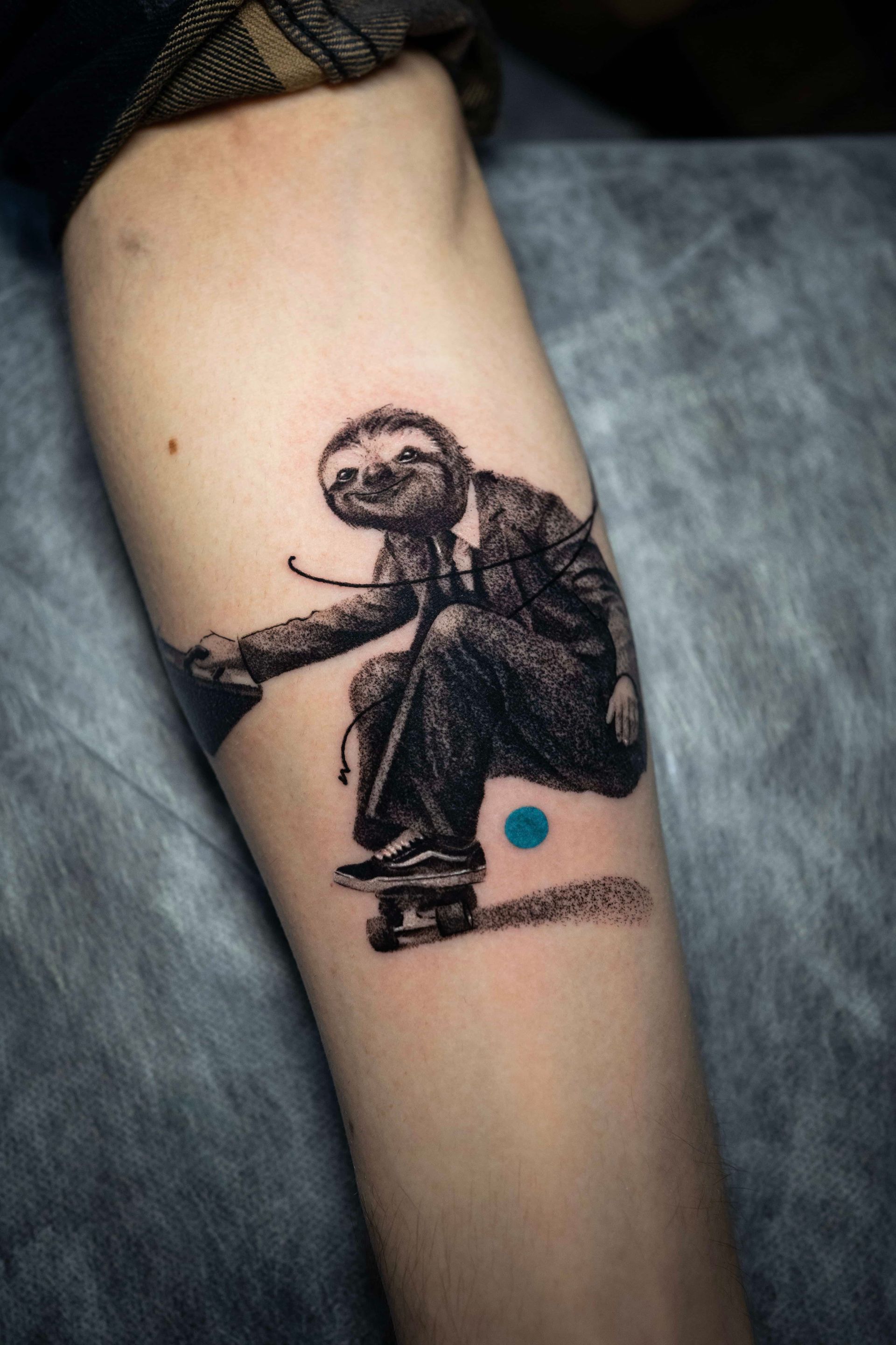 A forearm tattoo of a sloth wearing a suit and tie, riding a skateboard, with a small blue dot beside it.