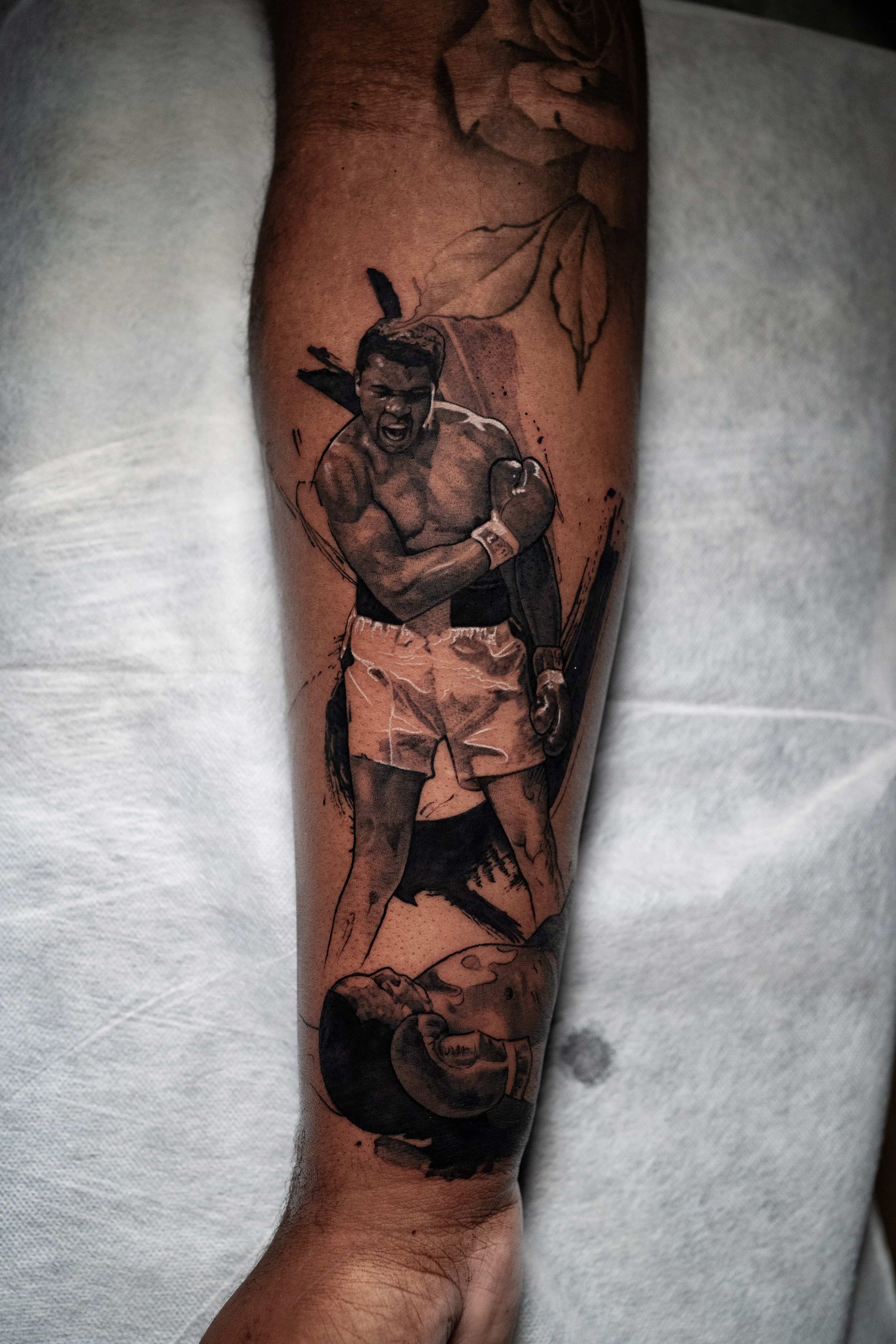 A black-and-grey tattoo on a forearm depicting a boxer in a fighting stance, surrounded by abstract, artistic shading.