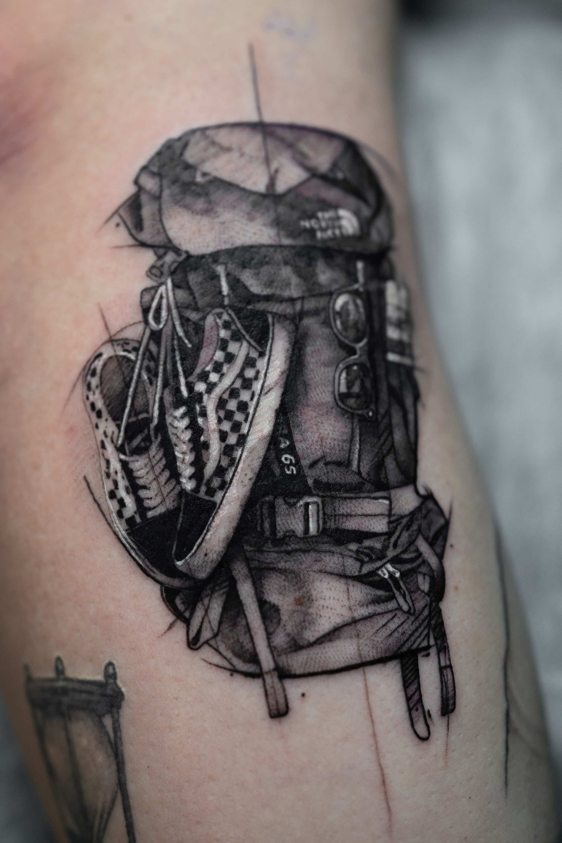 A black-and-grey tattoo of a hiking backpack with a pair of checkerboard Vans sneakers hanging from it.