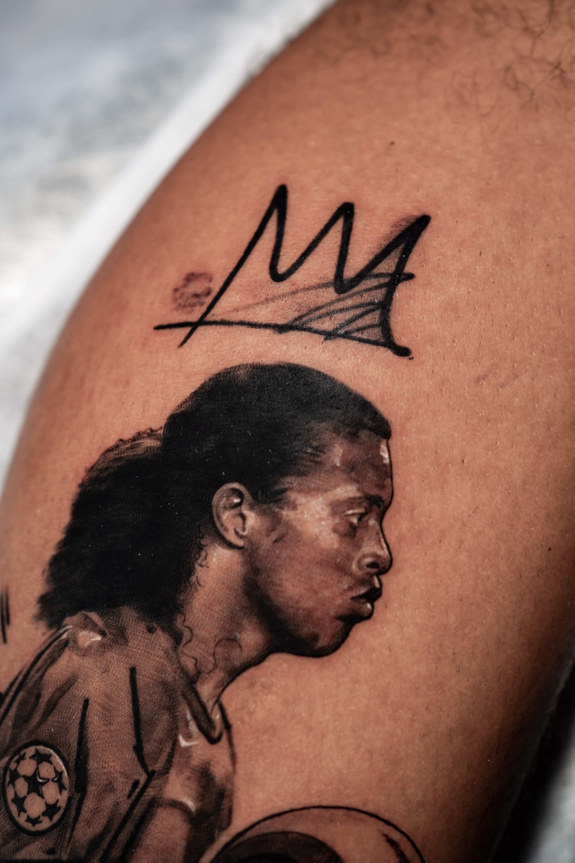 A tattoo on skin depicting Ronaldinho in profile, topped with a Basquiat-style crown symbol.
