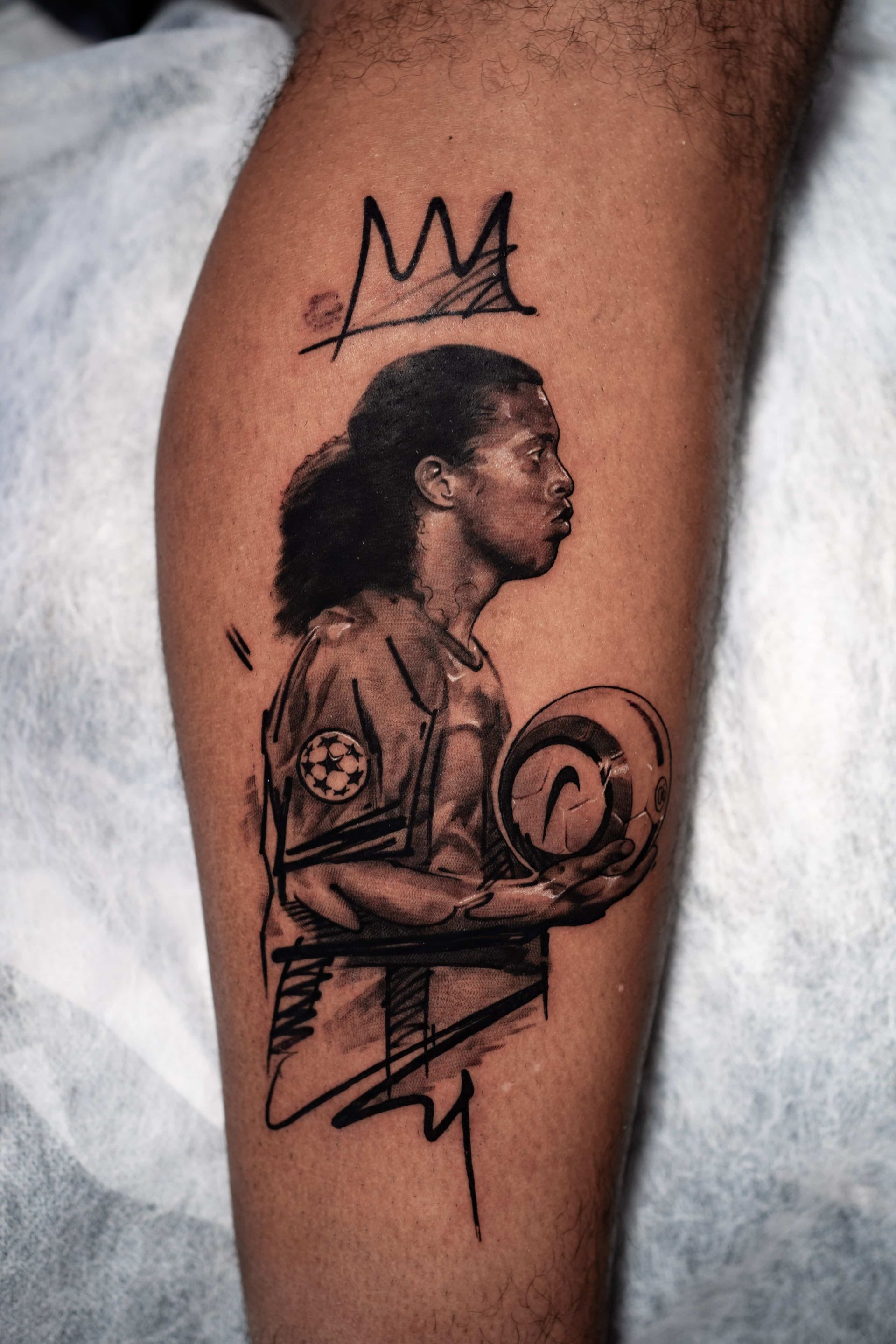 A tattoo of Ronaldinho in profile holding a soccer ball, with a crown drawn above his head in a sketch-like black ink style.
