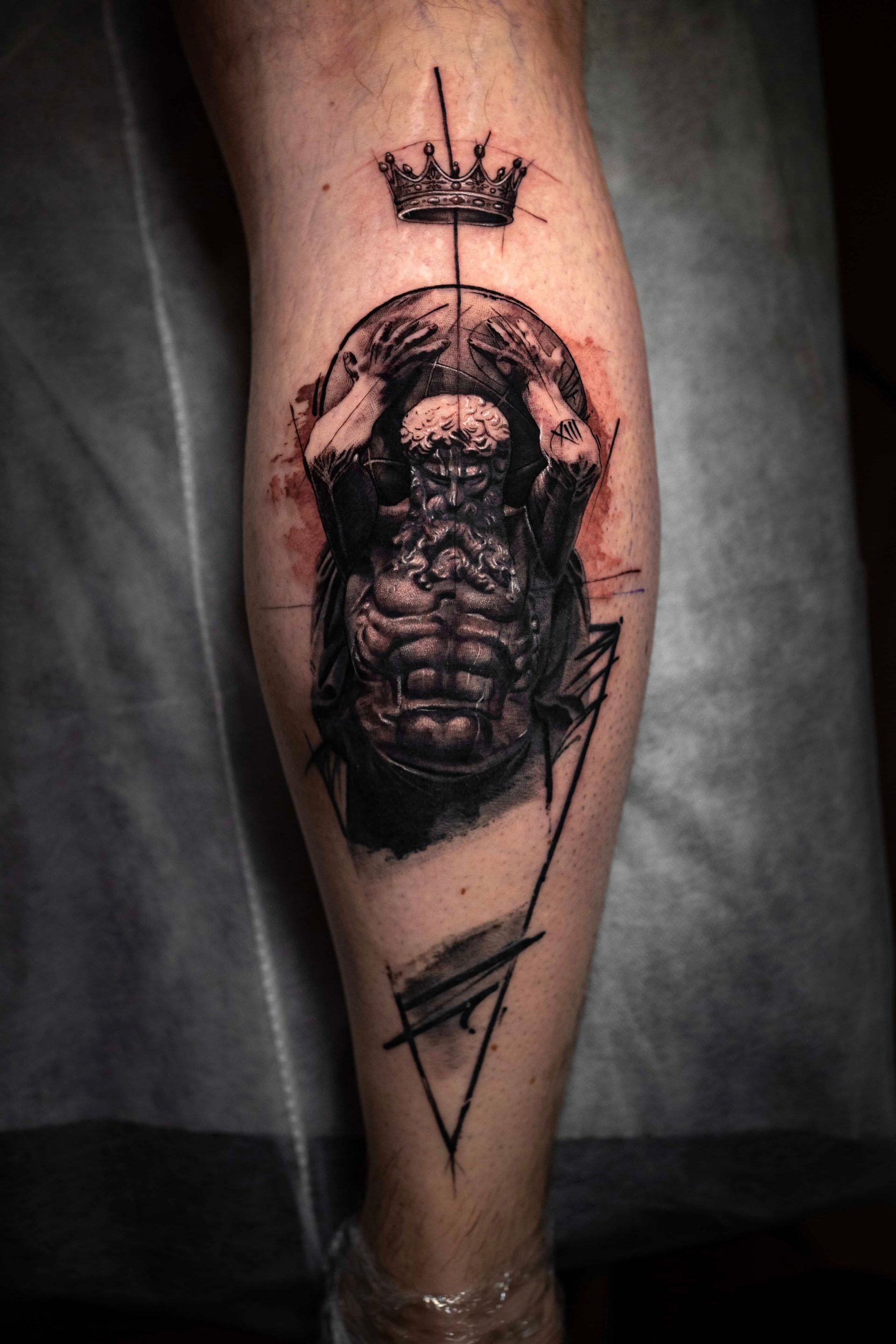 A calf tattoo featuring a dark, sketch-style figure holding a sphere aloft under a crown, accented by geometric lines.