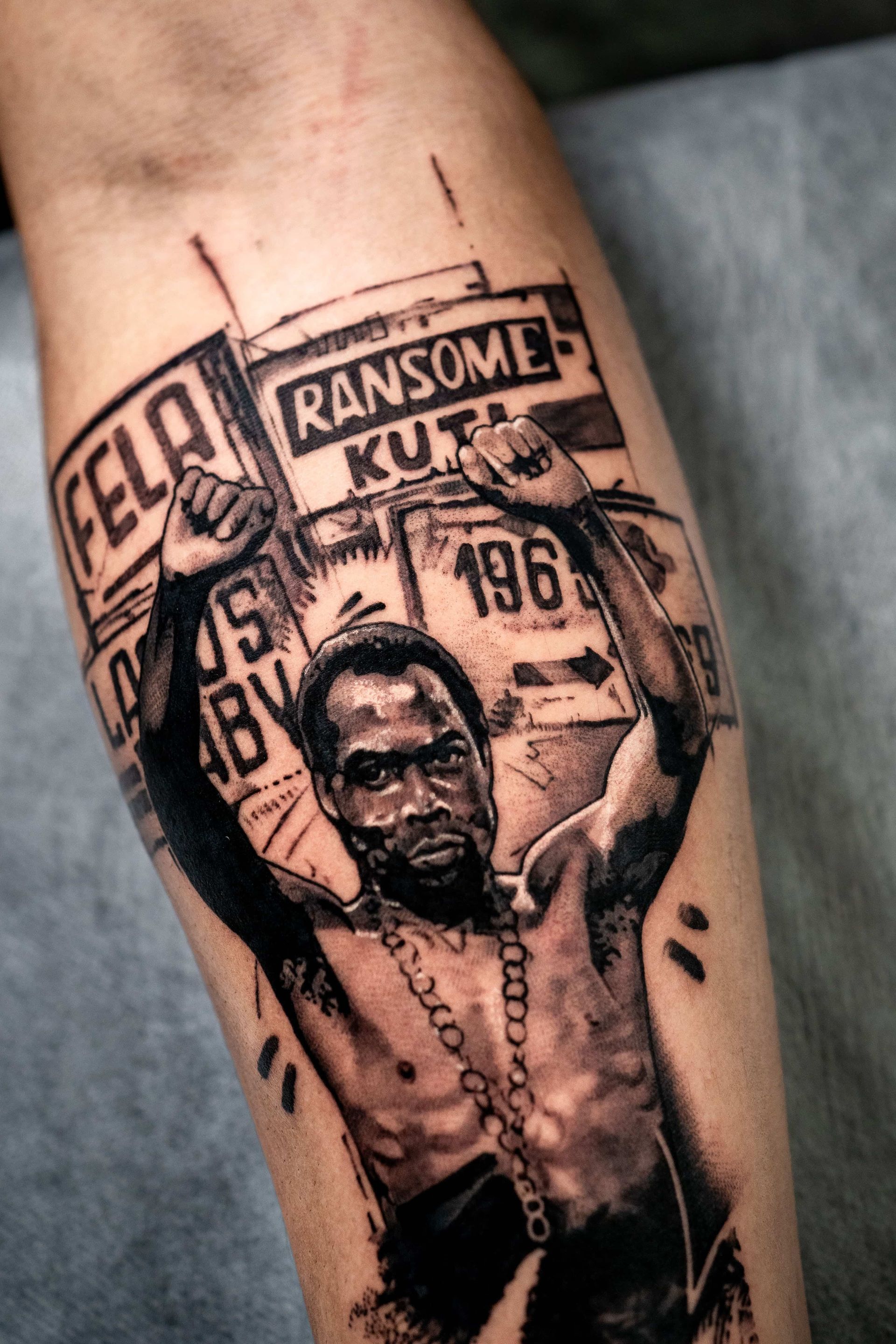 A black-and-grey tattoo of Fela Ransome Kuti with arms raised, set against a background of text and geometric shapes.