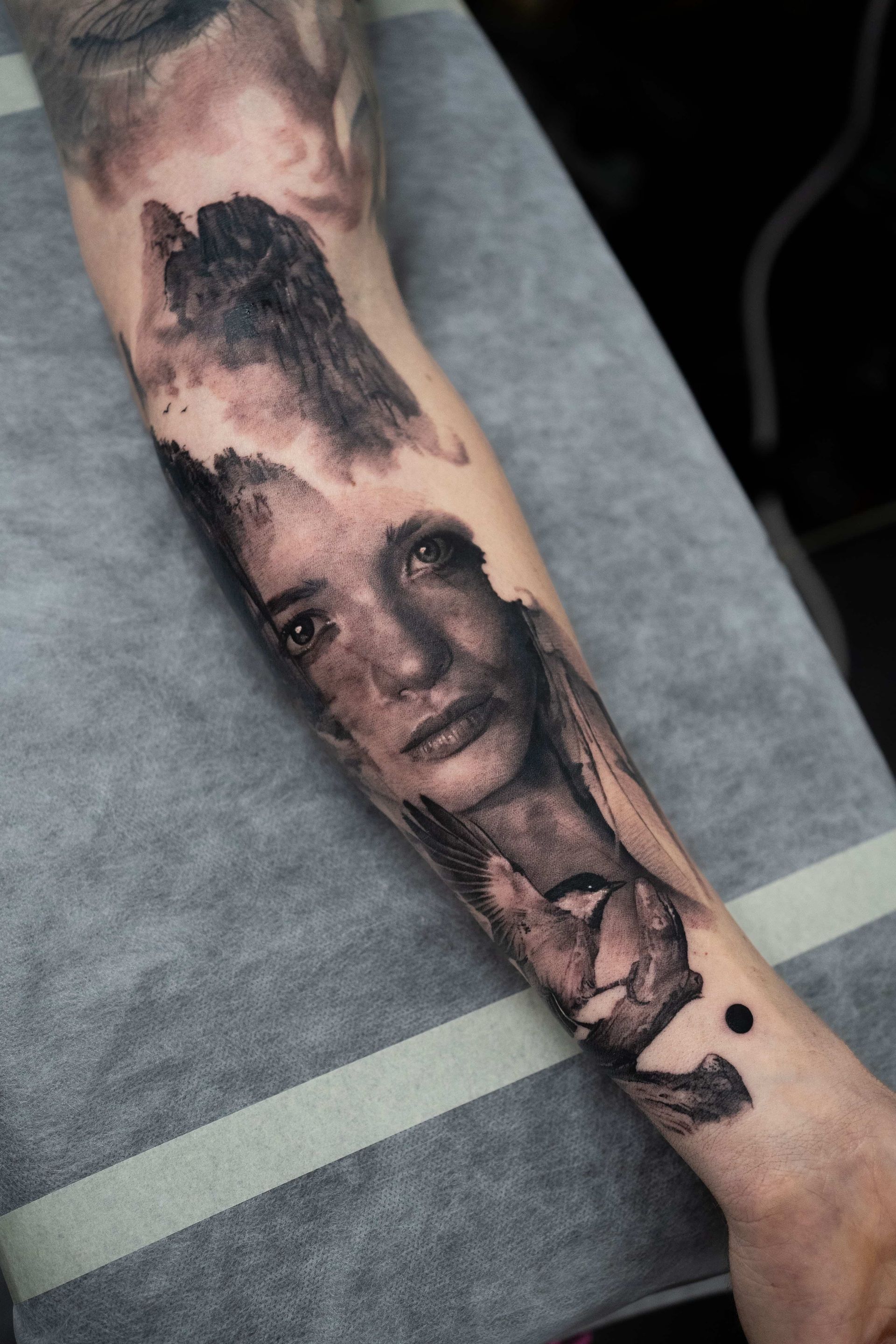 A black-and-grey tattoo on a forearm featuring a detailed portrait blended with dark, abstract, misty forest imagery.