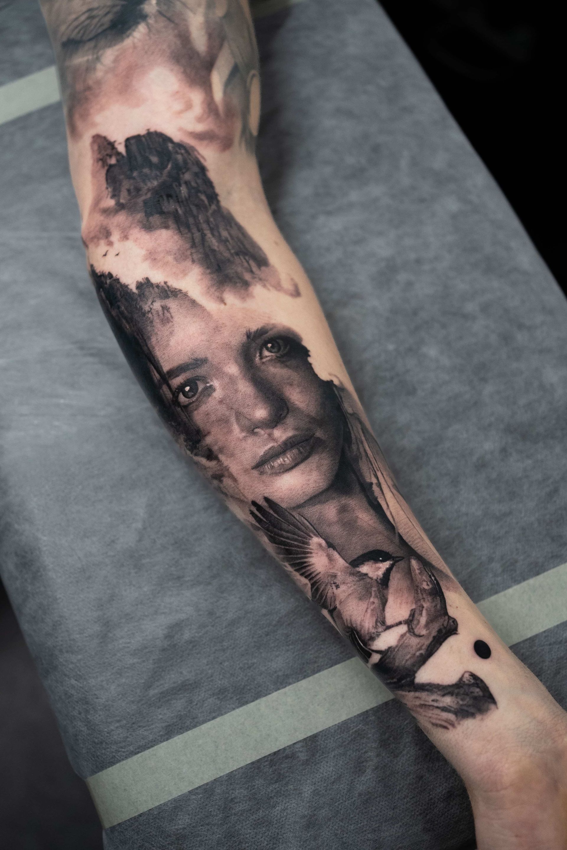 A forearm tattoo featuring a portrait integrated with scenes of a forest, mountains, and a bird in flight in black and gray.