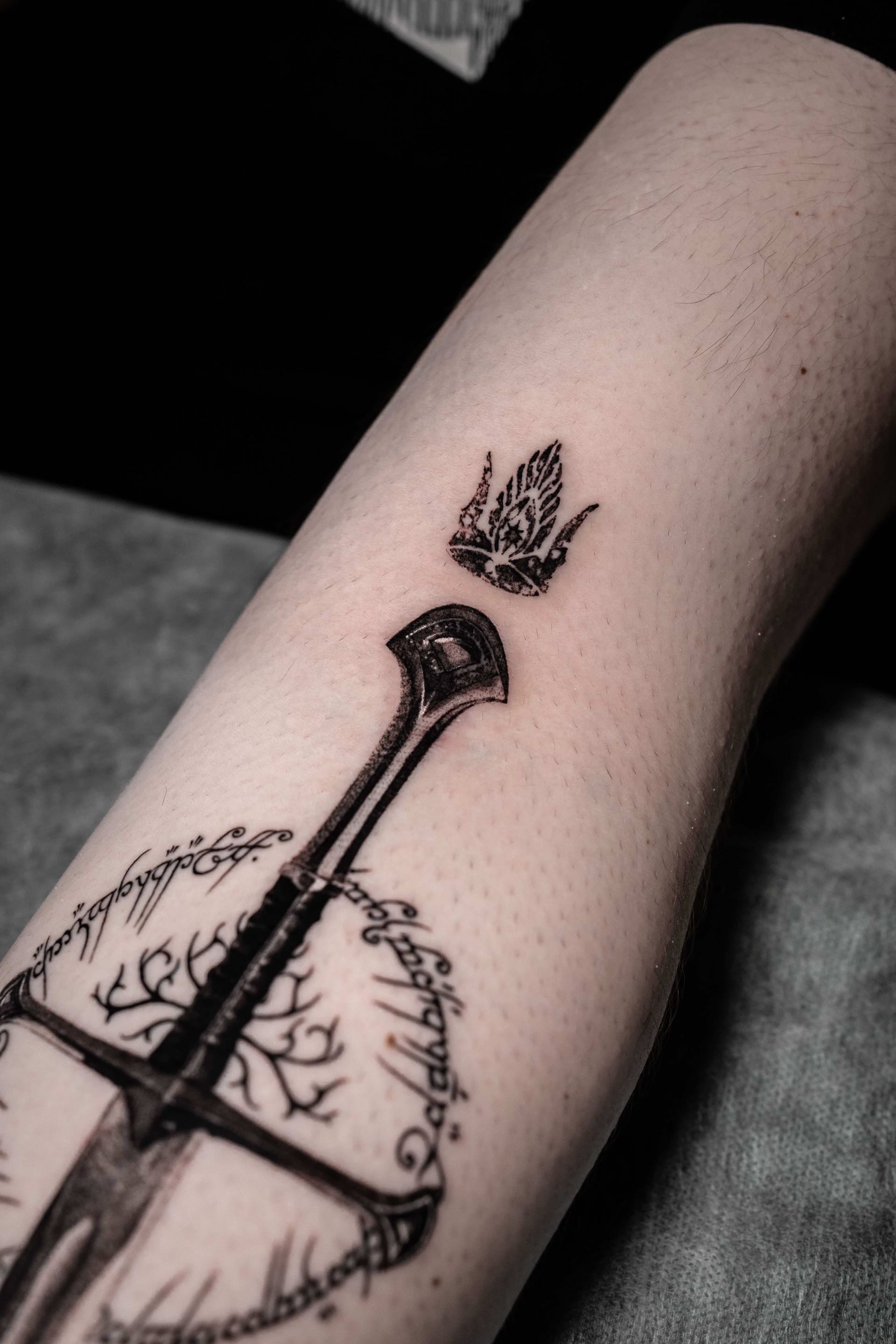 A black-ink tattoo on skin featuring a sword, the Tree of Gondor, Elvish script, and a crown above the hilt.