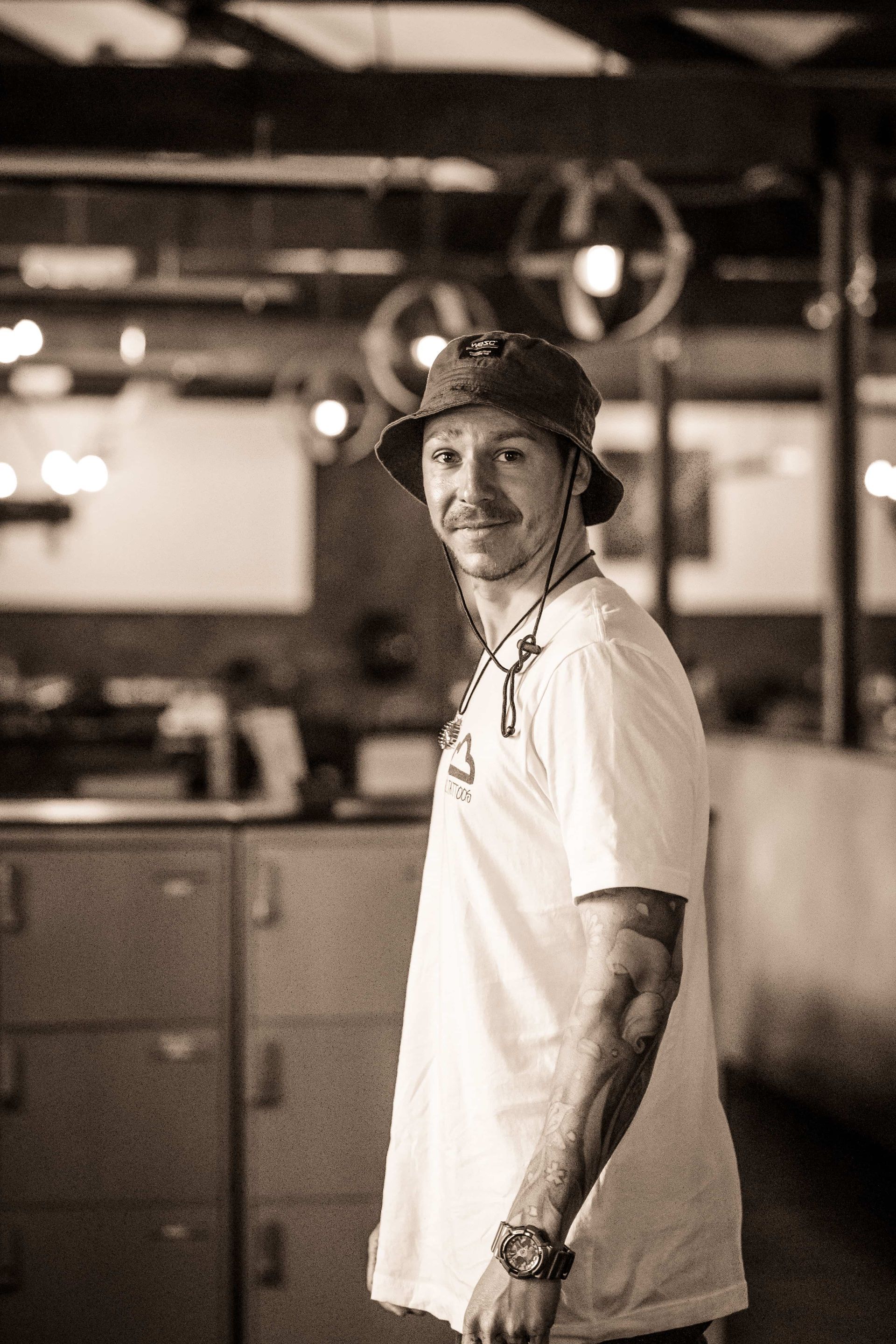 A person in a bucket hat and white t-shirt stands in a dimly lit space, looking over their shoulder at the camera.