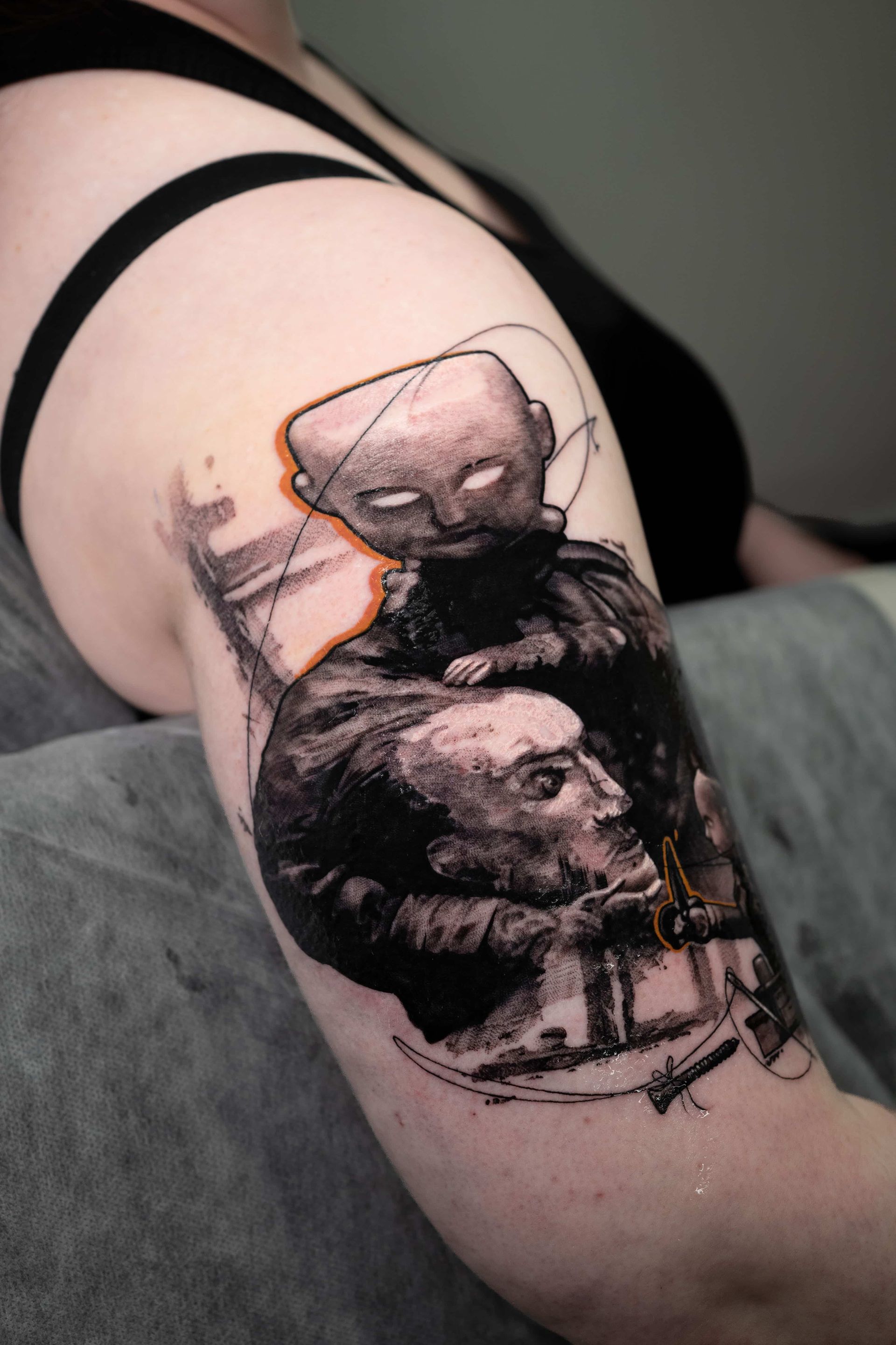 A tattoo on an upper arm featuring an etched-style illustration of two figures, with a bold orange line accenting a shape.