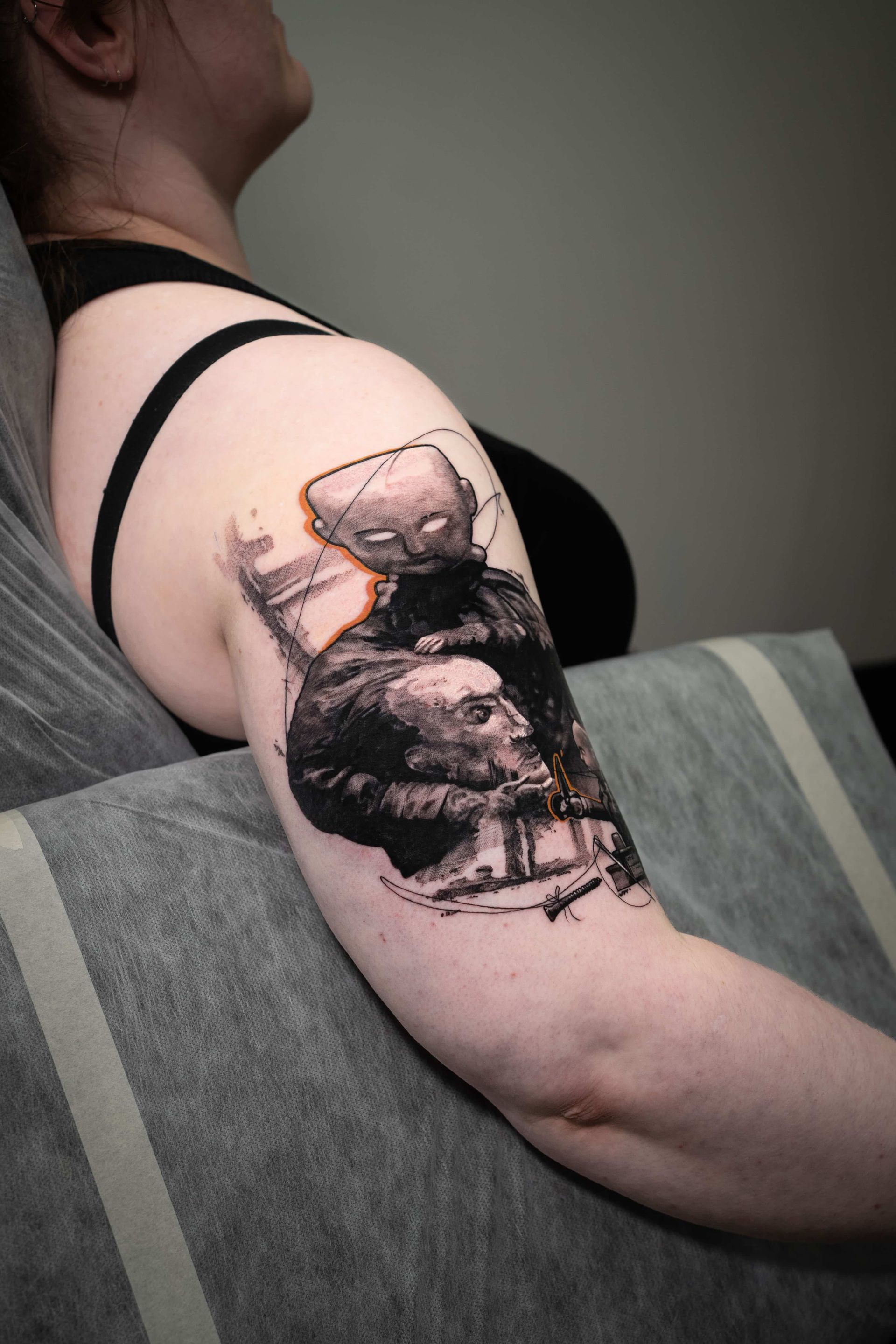 A person with a large, detailed black-and-white illustrative tattoo on their upper arm featuring abstract human figures.