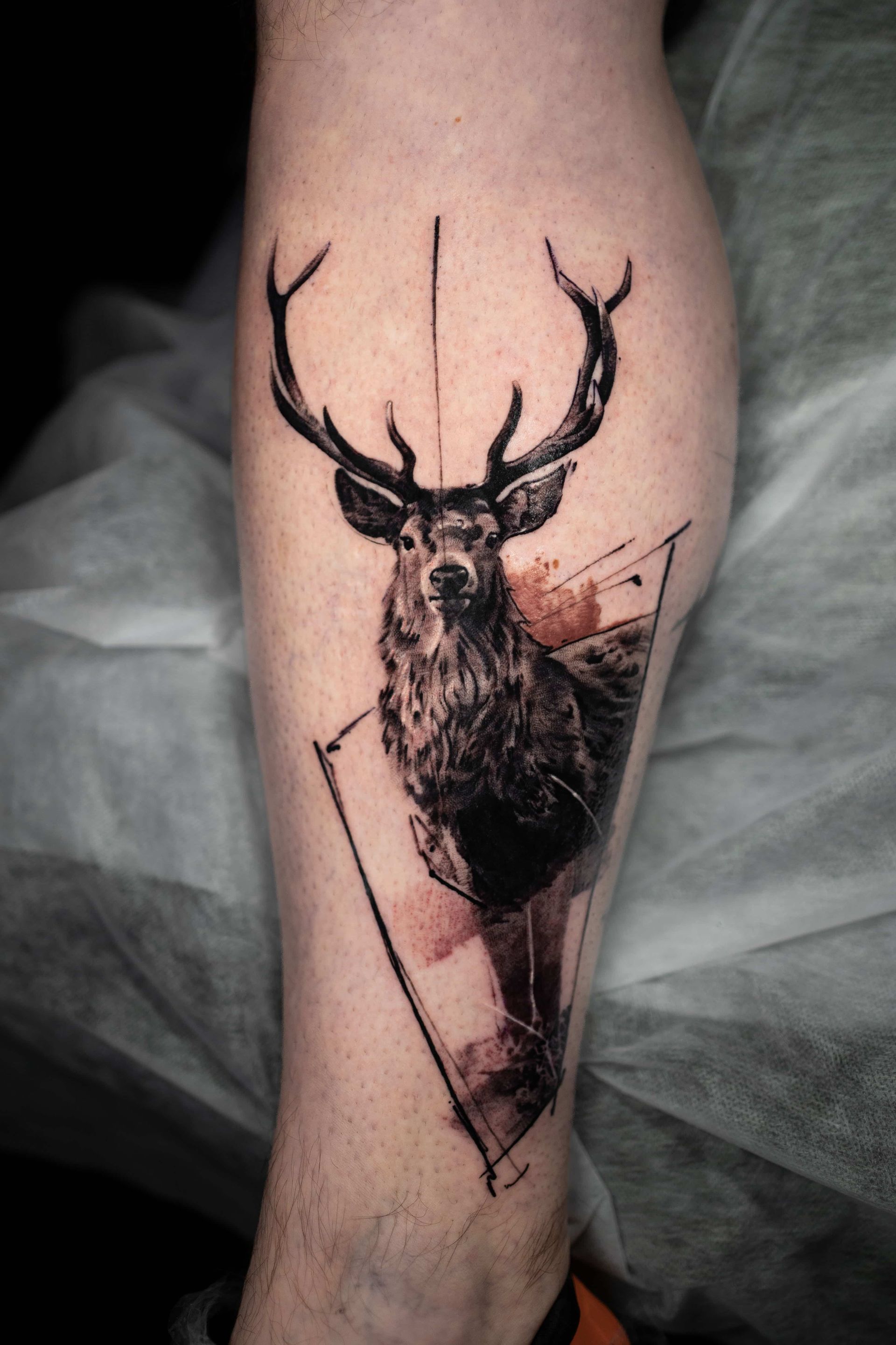A tattoo of a deer head with antlers, integrated into a geometric, watercolor-style design on a person's lower leg.
