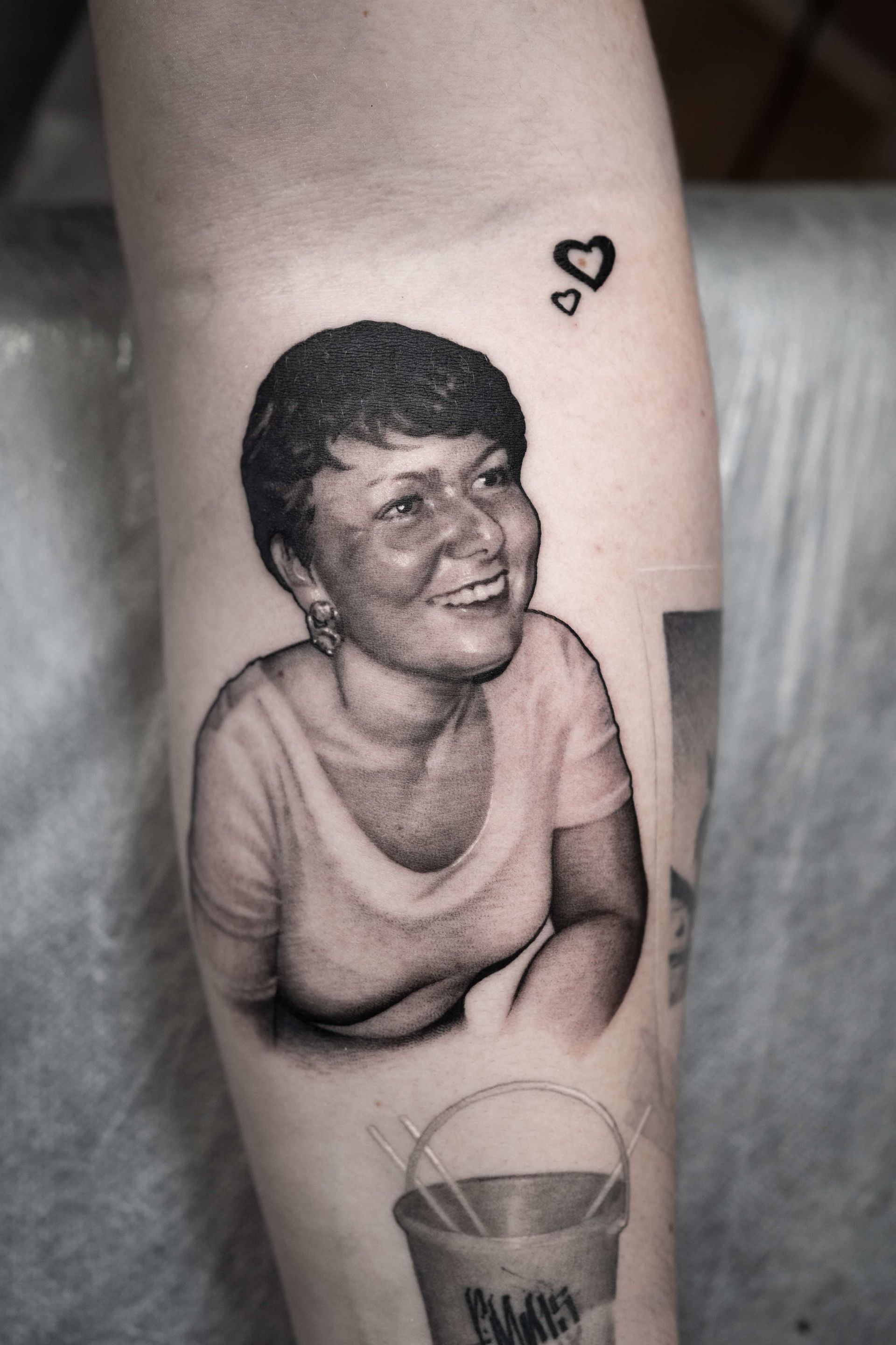 Portrait tattoo of a smiling person on an arm, with two small heart outlines above and a bucket tattoo below.