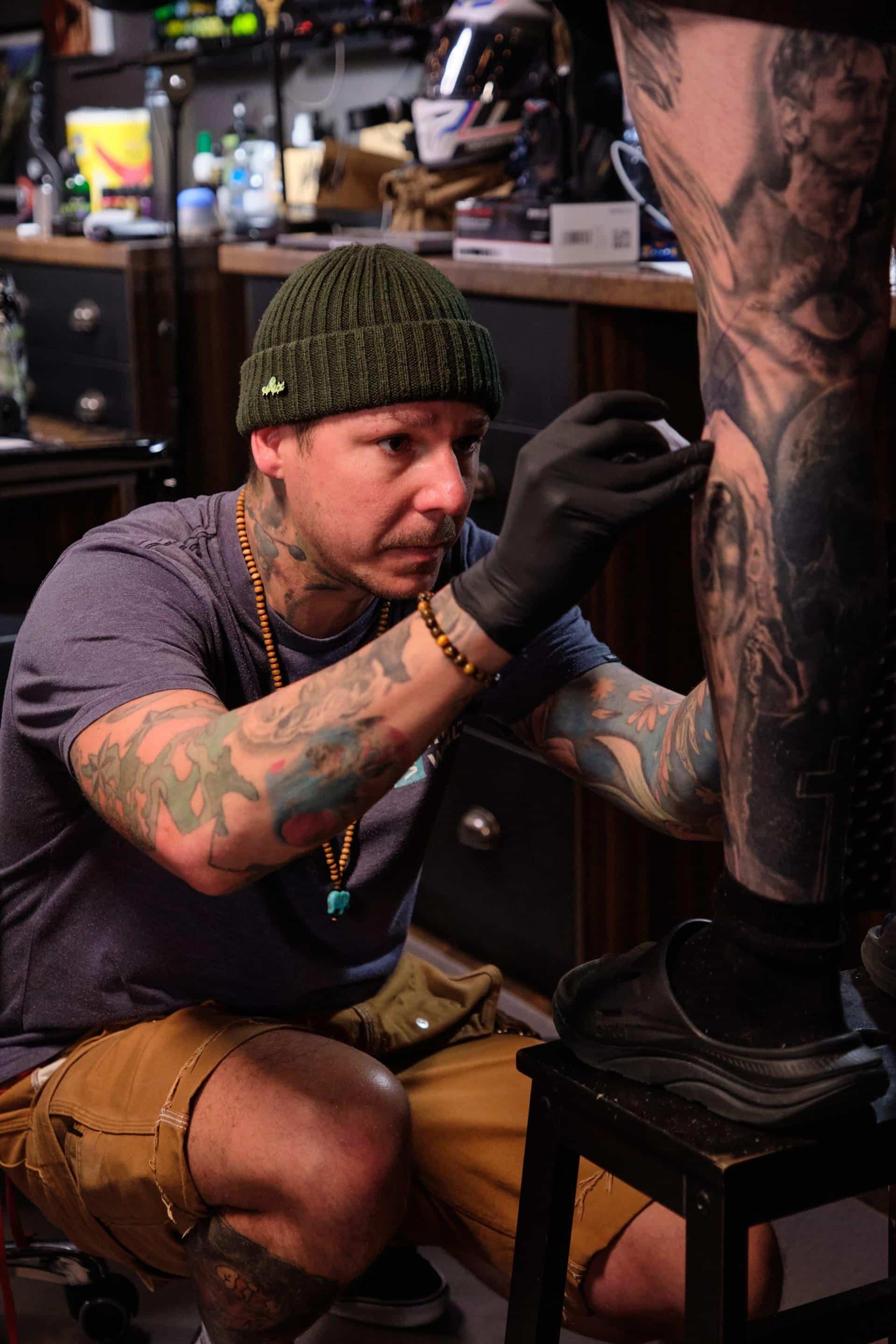 A tattoo artist in a green beanie and black gloves focuses intently on tattooing a detailed design on a client's leg.