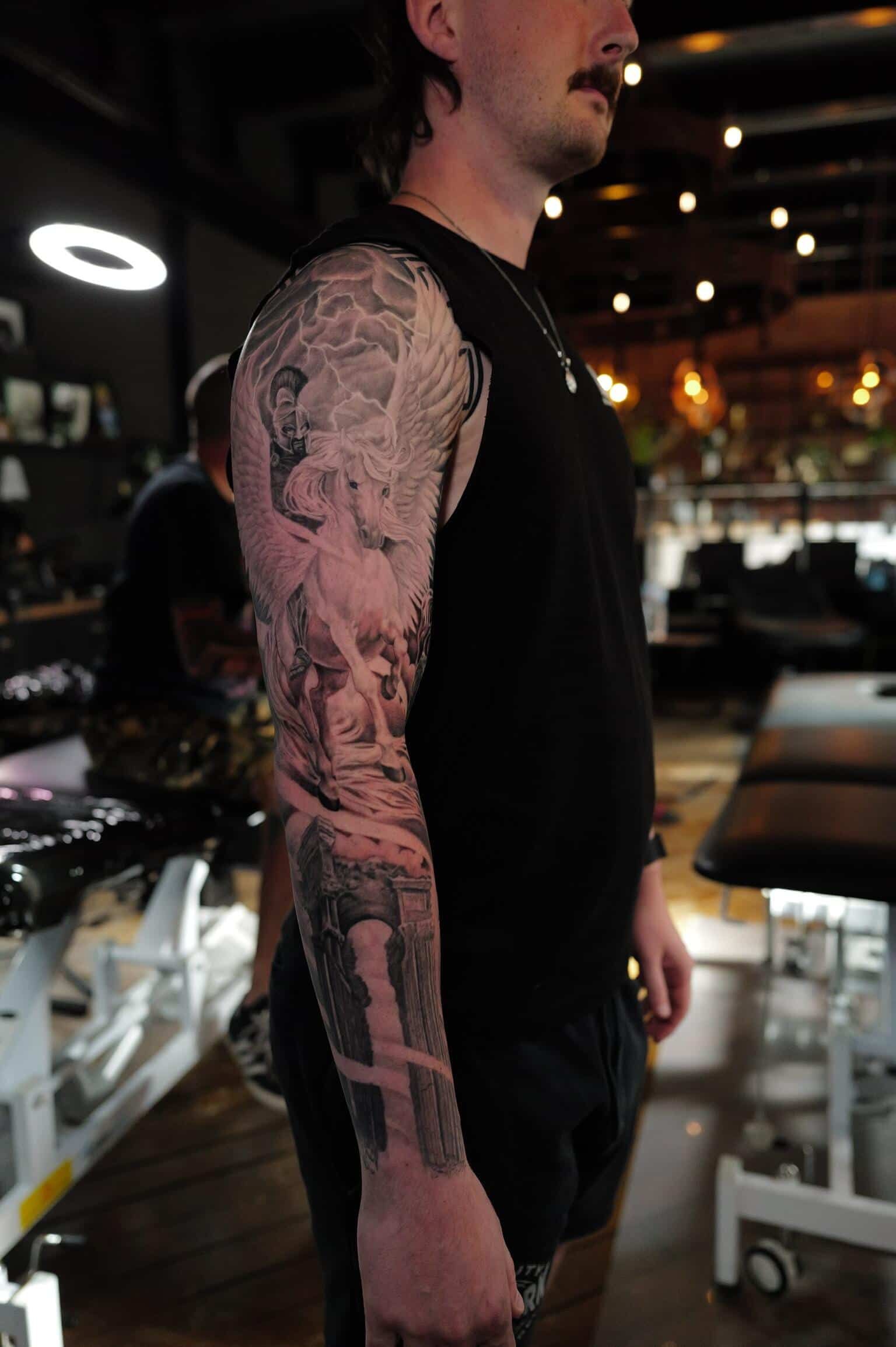 A person in a tattoo studio shows a finished dark-themed illustrative sleeve tattoo covering their entire arm.